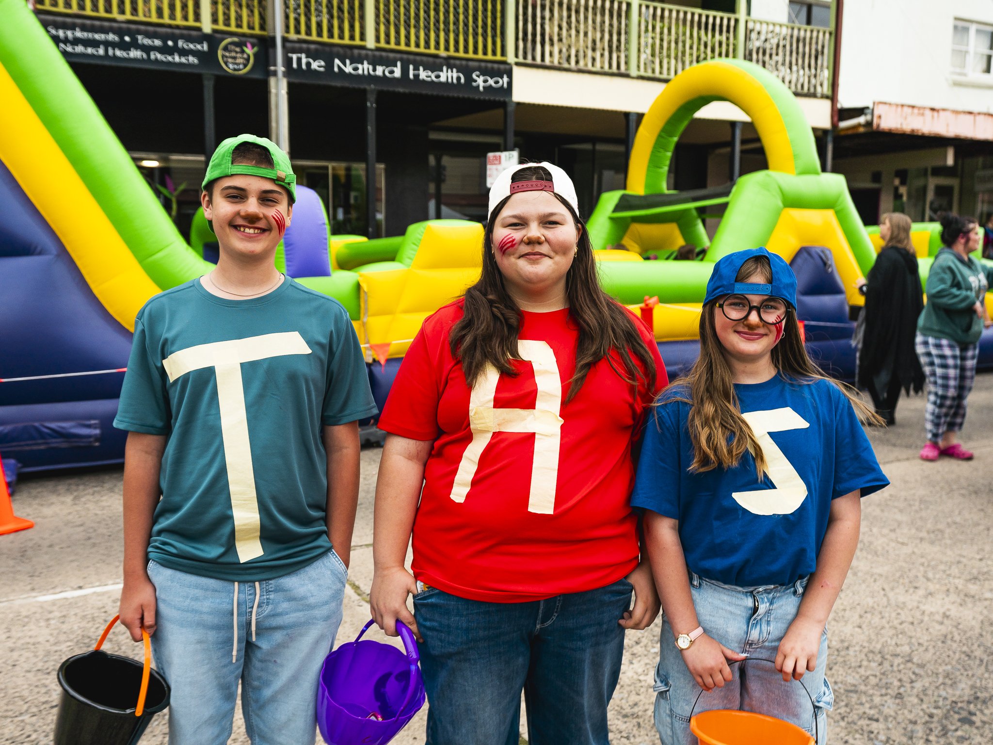 Three girls standing in front of a colorful inflatable bounce house, wearing shirts with the numbers 1, 5, and 3 on them, with face paint and hats, holding buckets for a celebration or event.