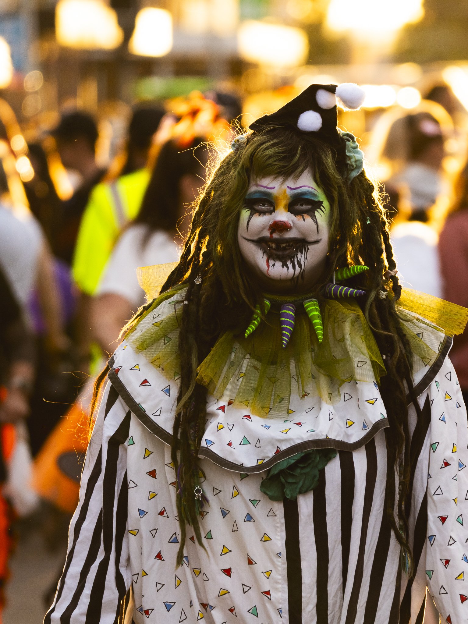 Person dressed in clown costume with makeup, dreadlocks, and colorful costume at a festive event.