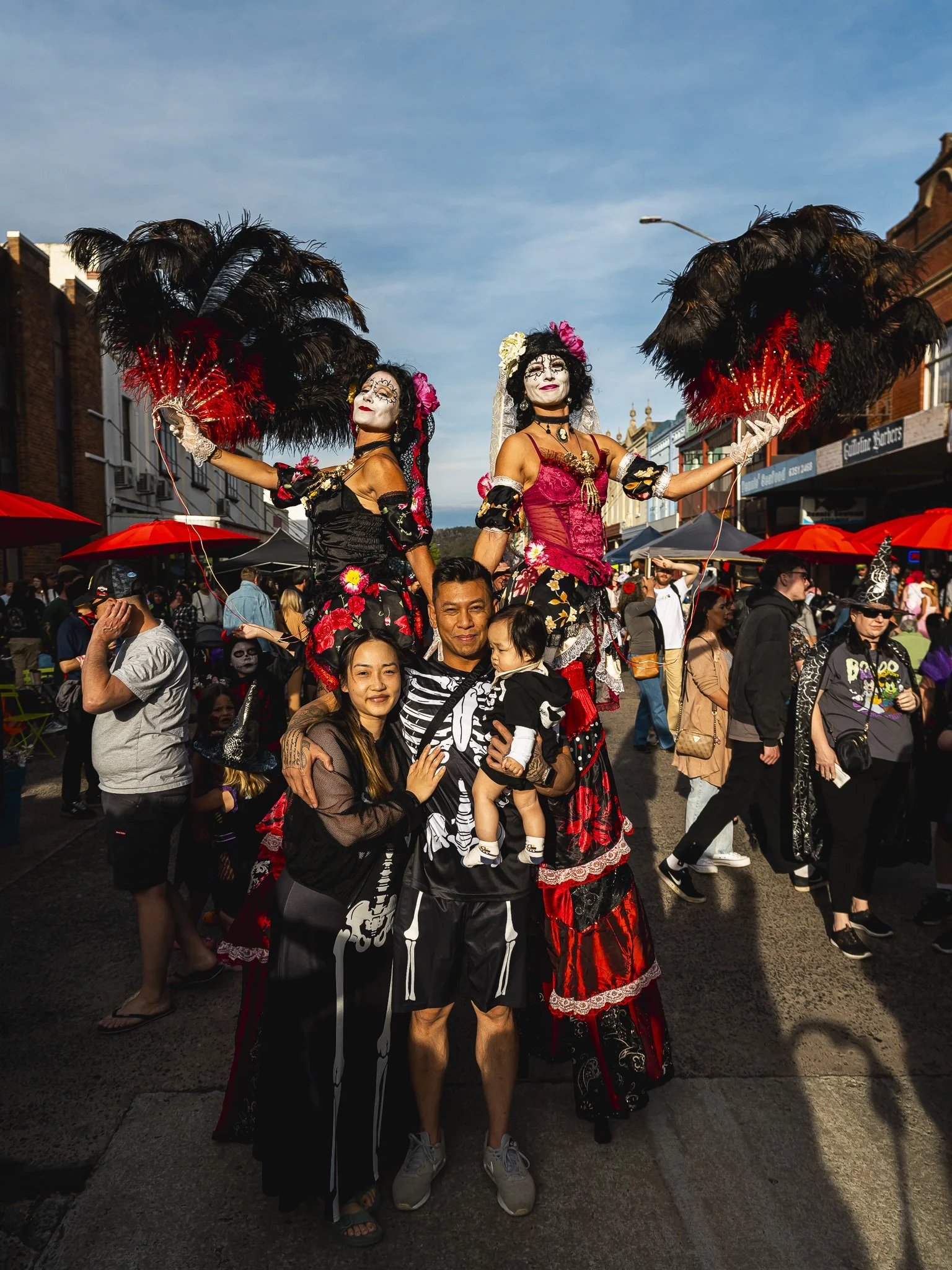 Family celebrating Dia de los Muertos, with two performers on stilts dressed in colorful, traditional costumes, surrounded by a crowd in a lively street.