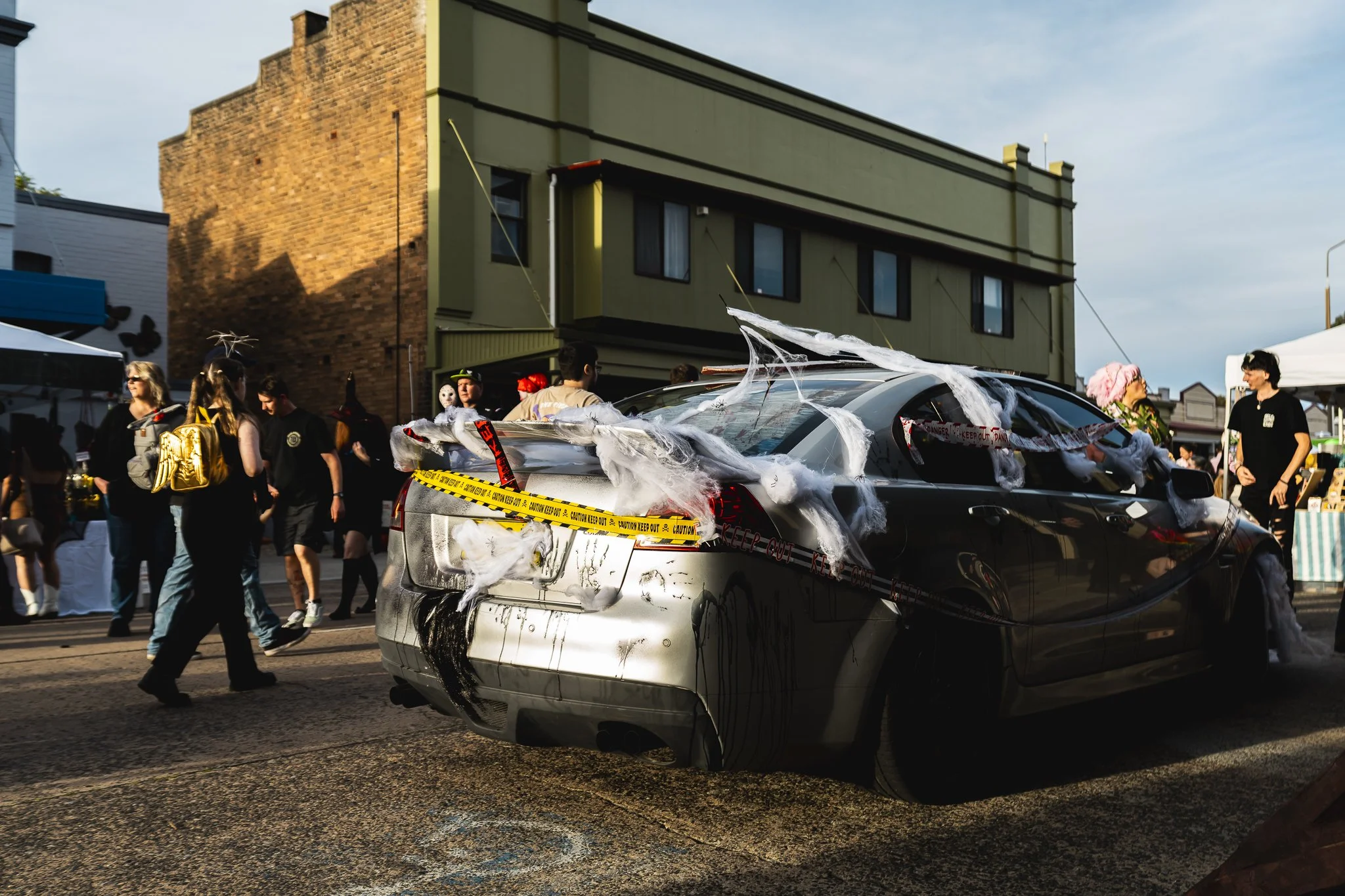 A black car decorated with fake spider webs and caution tape at a Halloween event, with people in costumes walking nearby on a city street.