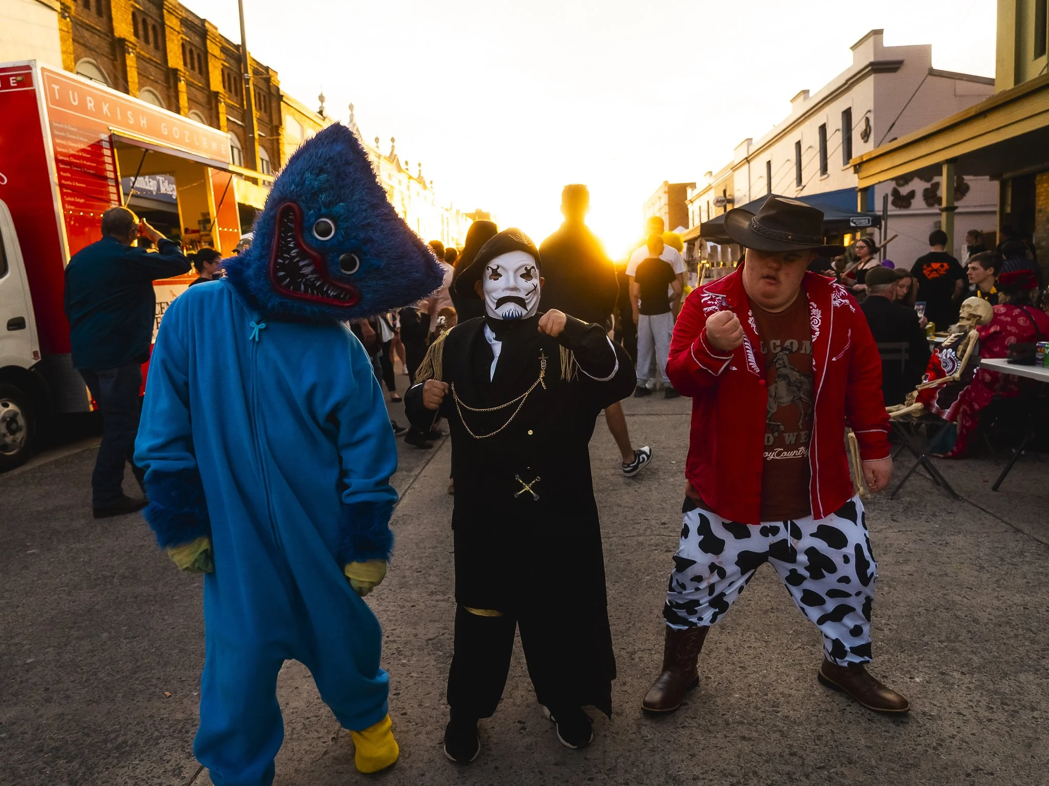 Three people in costumes at a street festival during sunset; one in a large blue shark costume, one in a black and white clown mask with a black outfit, and one in a cowboy hat, red jacket, and cow-print pants.