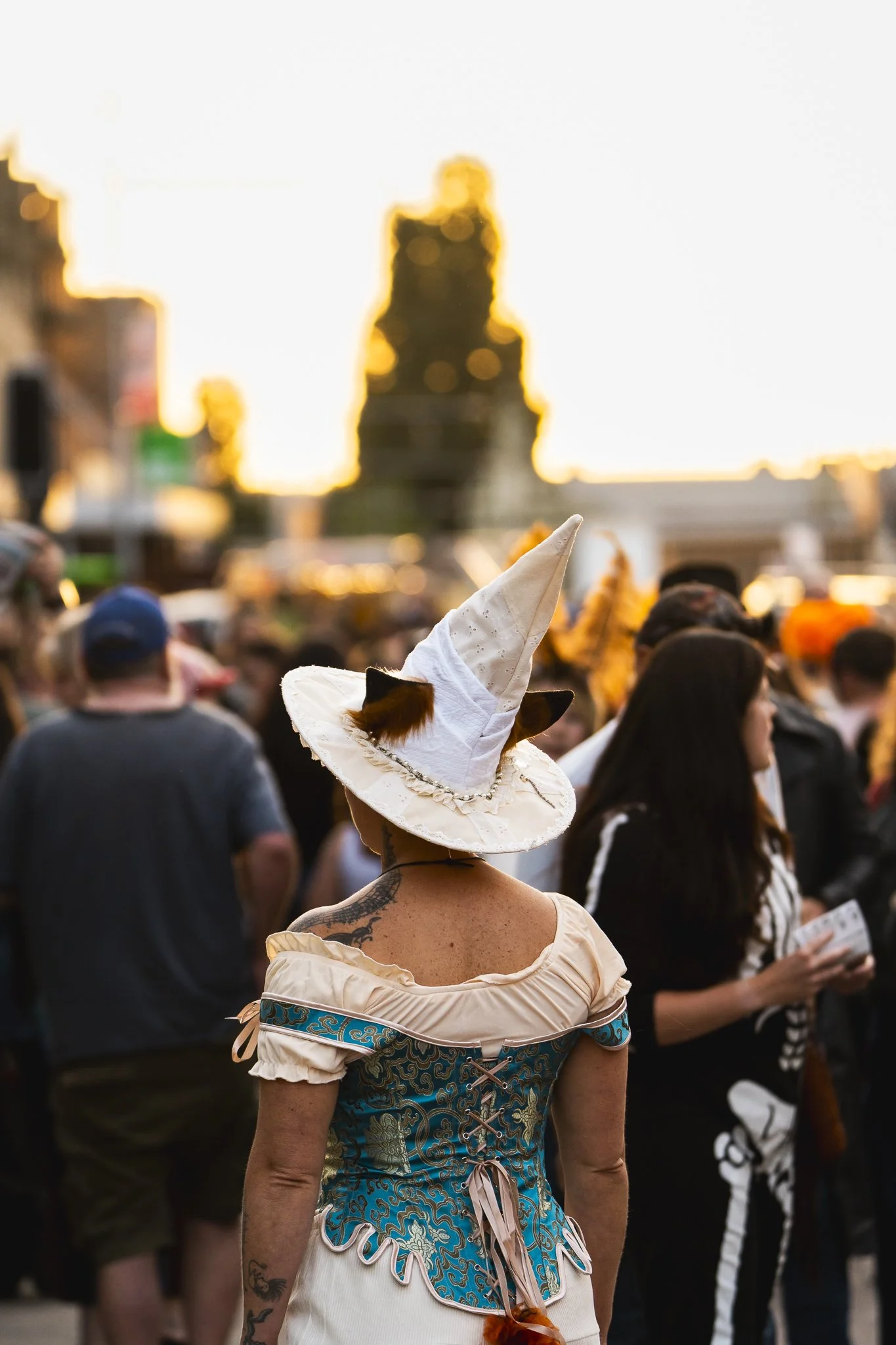 A person dressed as a witch with a tall, pointed hat and a corset-style dress standing amidst a crowd outdoors during sunset.