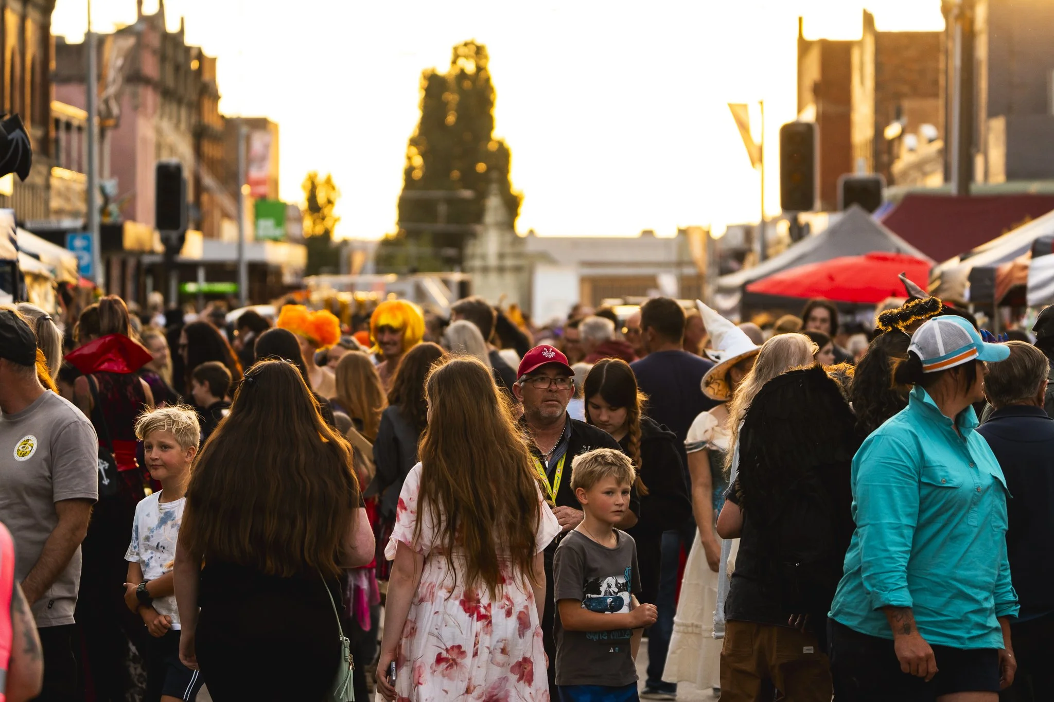 Crowd of people at an outdoor festival or street fair in the evening with tents and booths in the background.