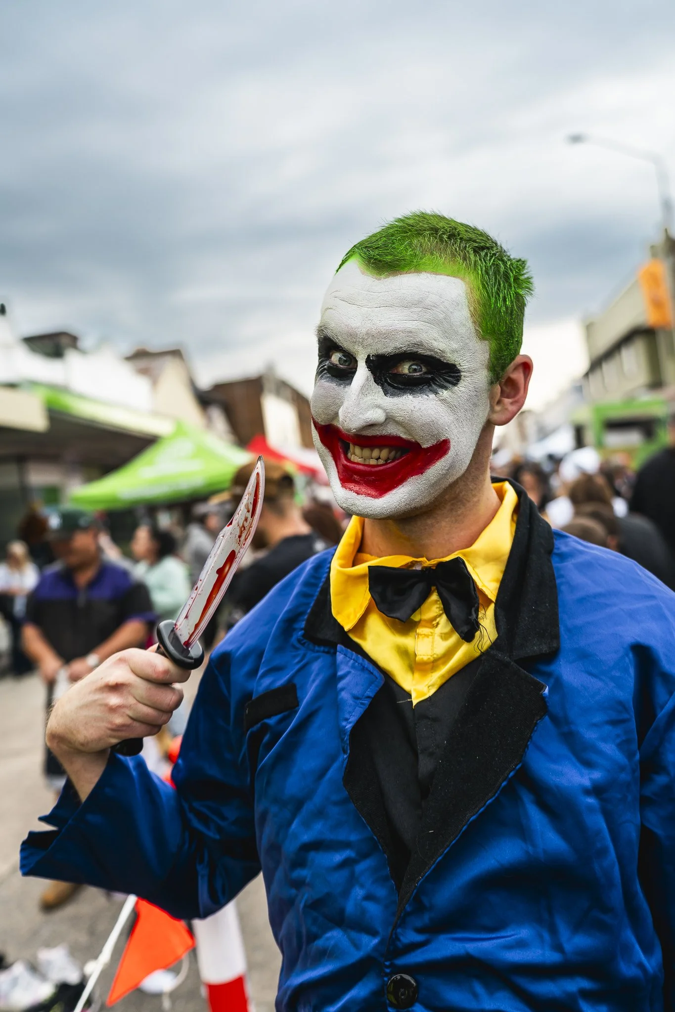 Person dressed as Joker with green hair, white face paint, black eye makeup, and red lipstick, holding a bloody toy knife, at an outdoor event with a crowd and vendor stalls in the background.