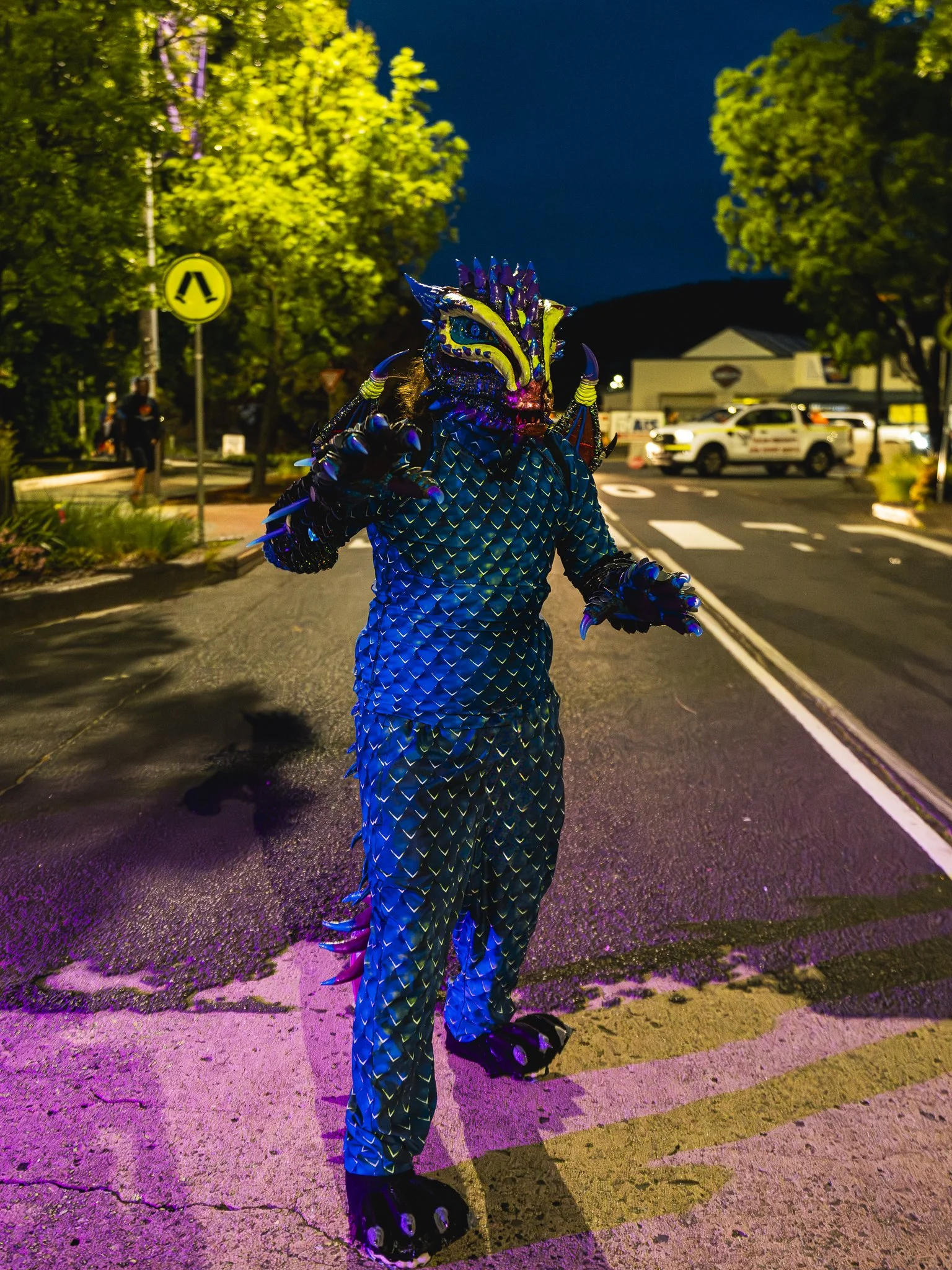 A person dressed in a colorful dragon costume with a detailed, scaled design, standing on a city street at night, illuminated by streetlights and cars in the background.