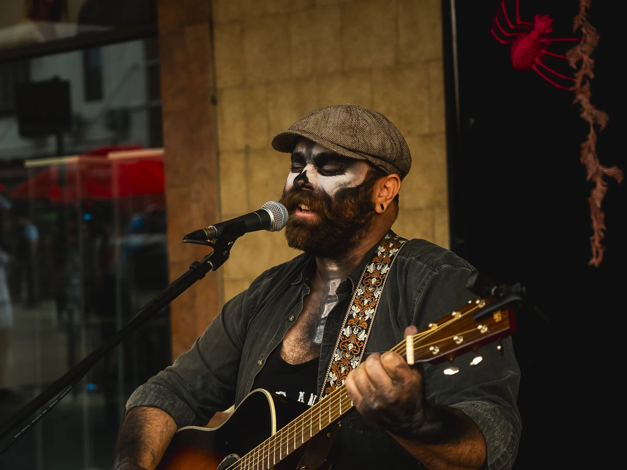 A man with painted face, beard, and mustache singing into a microphone while playing an acoustic guitar, wearing a flat cap and a dark shirt.