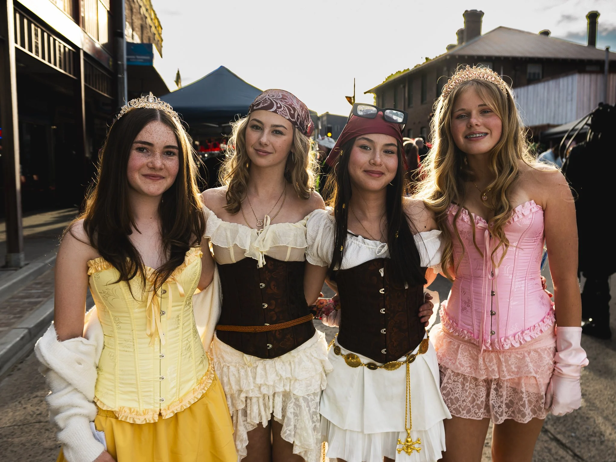 Four young women dressed in colorful, elaborate costumes with tiaras and accessories, smiling outdoors during a festival or event.