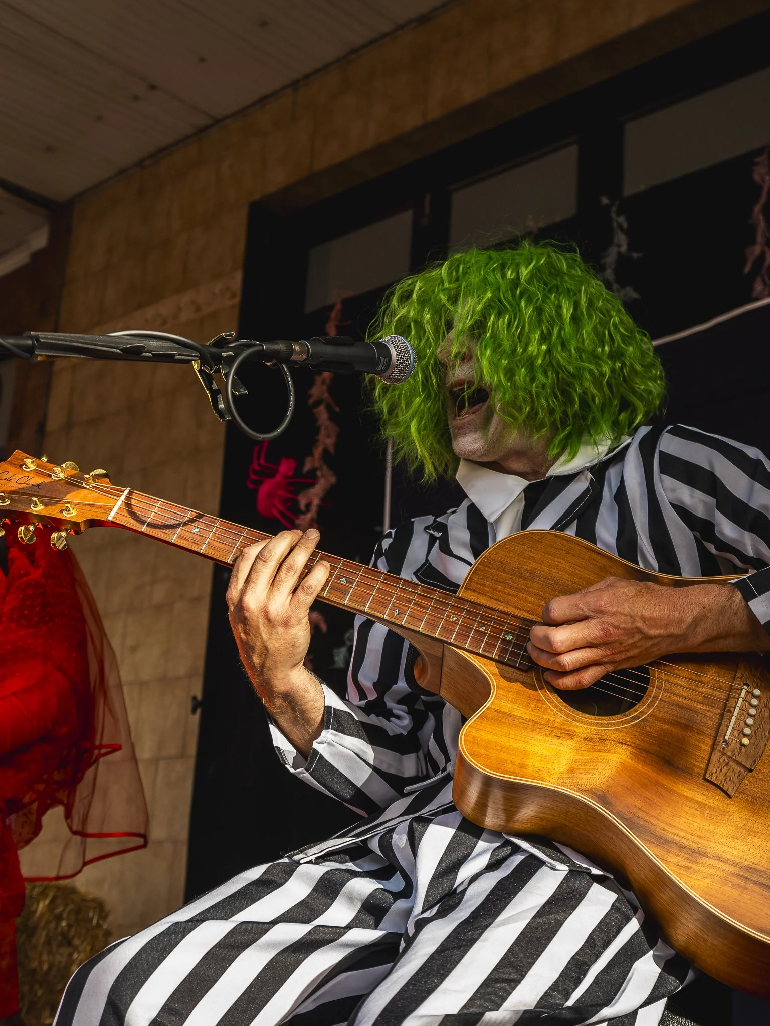 A man with bright green curly hair performing with an acoustic guitar on stage, singing into a microphone, wearing black and white striped pajamas.