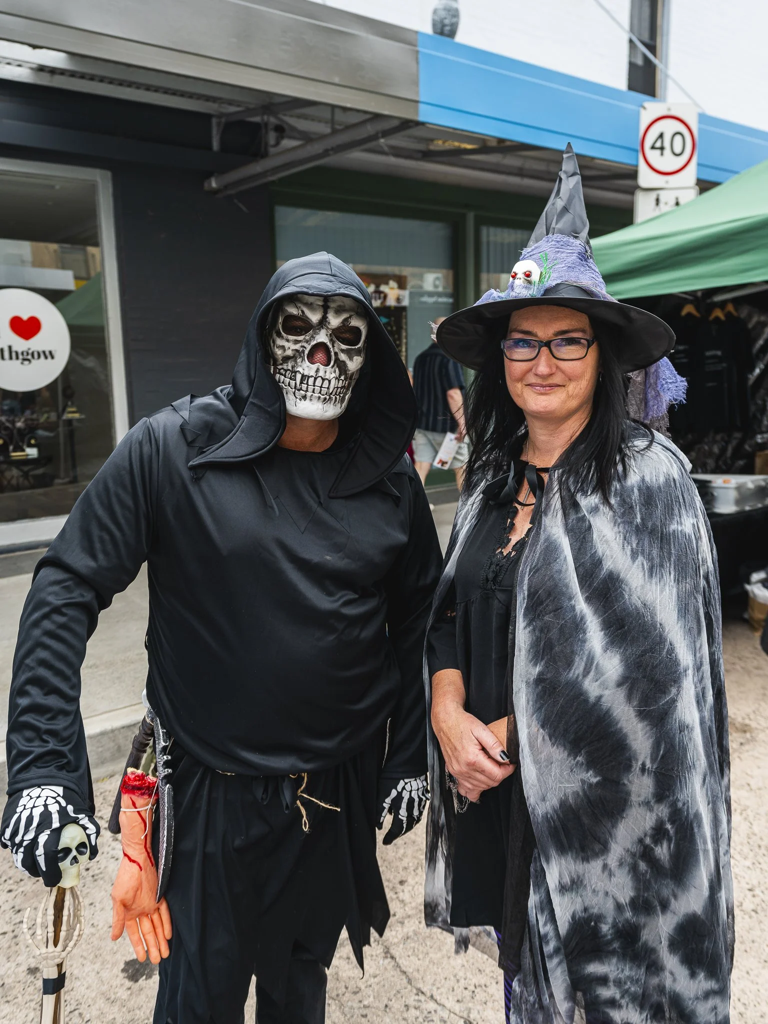Two people dressed in Halloween costumes standing outdoors in front of a building and a tent. One person is wearing a skeleton mask and black hooded outfit, and the other is dressed as a witch with a pointed hat and a gray and black cape.