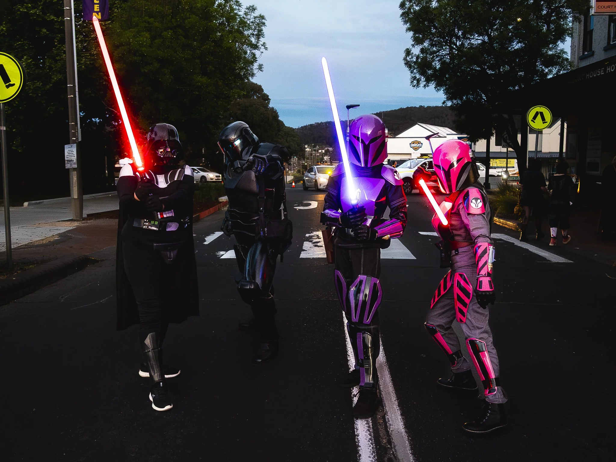 Four people dressed as Star Wars characters, including Darth Vader, Mandalorian, and Bounty Hunter, holding colorful glowing toy lightsabers standing on a city street at dusk.