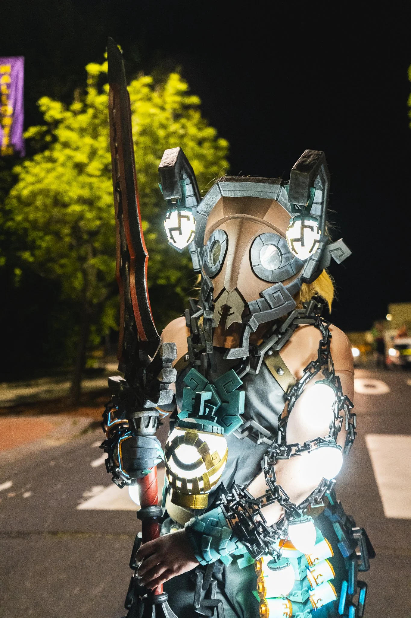 A person in a detailed, futuristic costume with glowing lights, chains, and a mask resembling a robotic animal, standing outdoors at night.