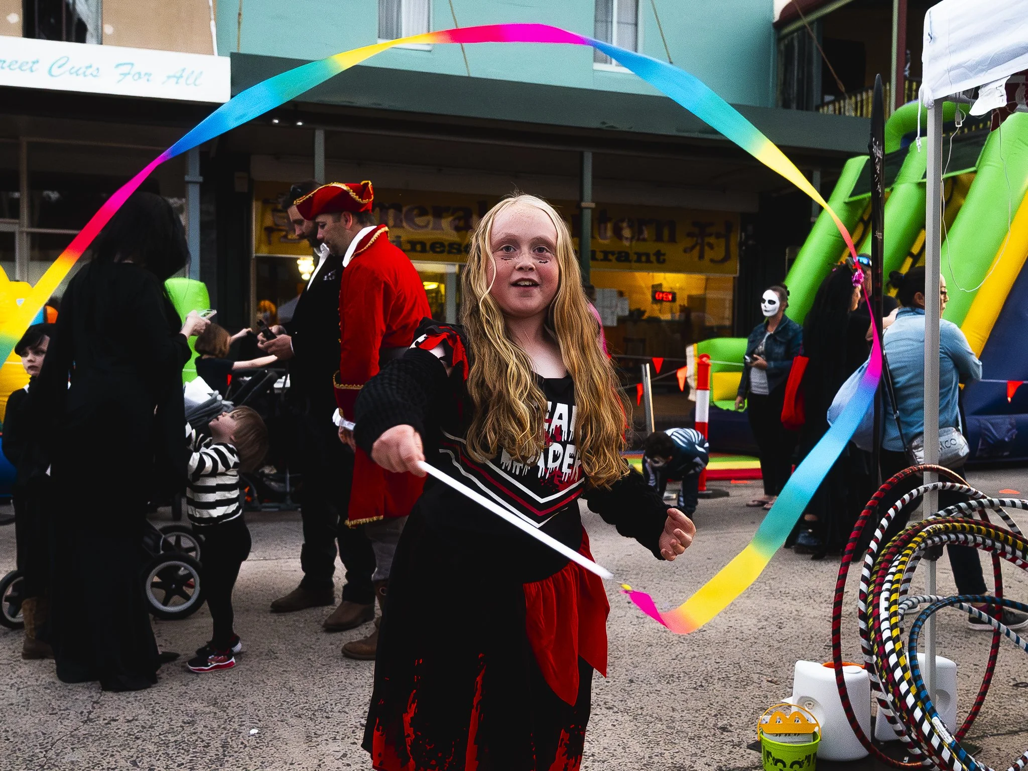 Young girl with long red hair dressed in a costume holding a stick, participating in a festive outdoor event with people in costumes, an inflatable slide, and colorful decorations.
