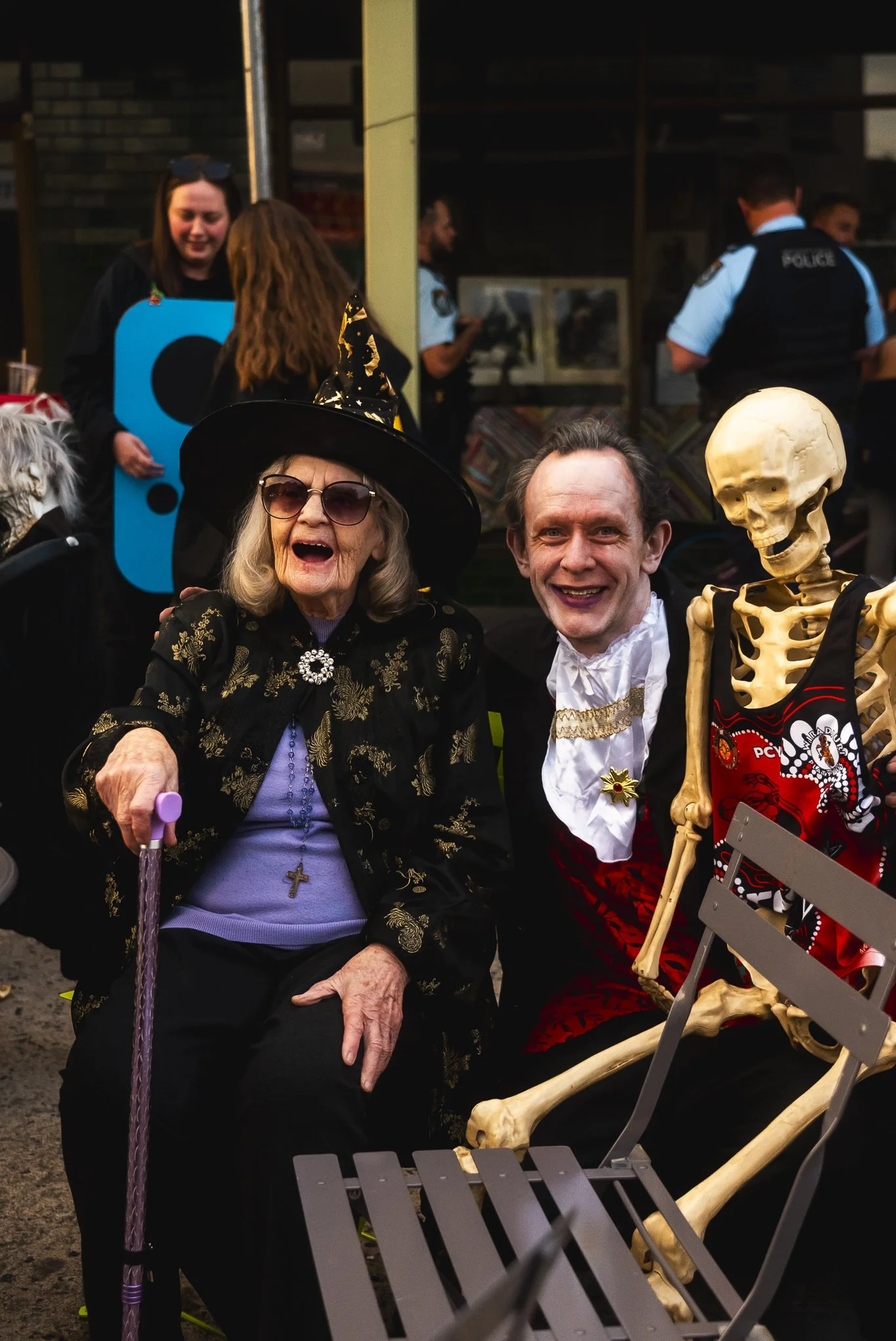 An elderly woman dressed as a witch with glasses, a cane, and a cross necklace sitting next to a man in a costume, and a skeleton wearing a hockey jersey, with people and police in the background.