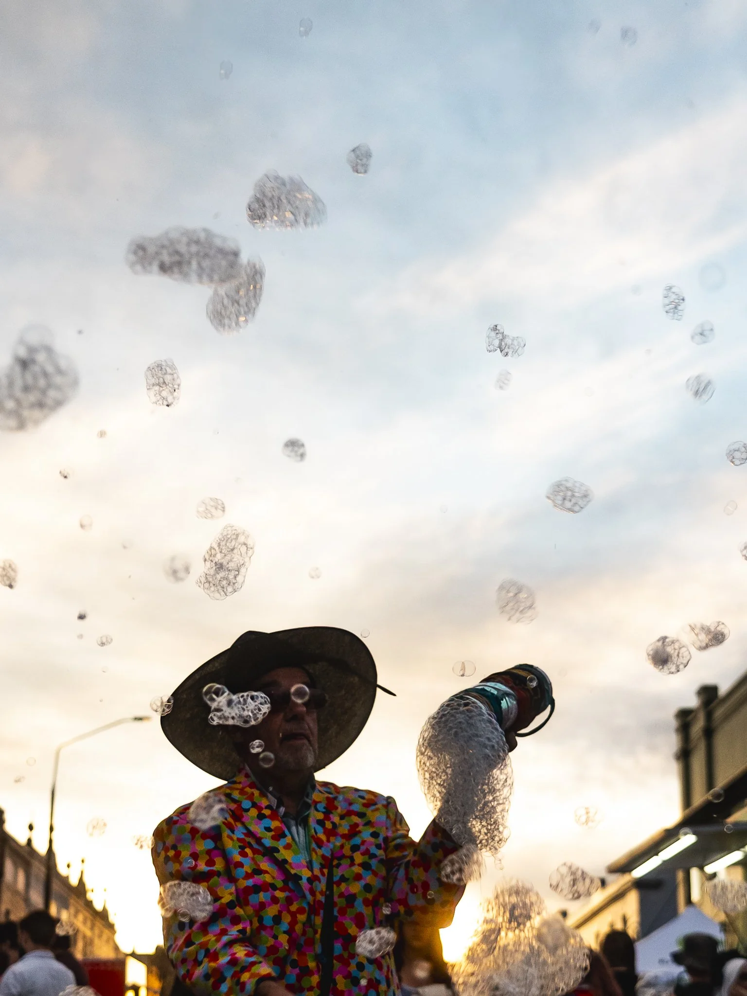 A person wearing a colorful polka-dot jacket, hat, and sunglasses selling soap bubbles at sunset during an outdoor event.