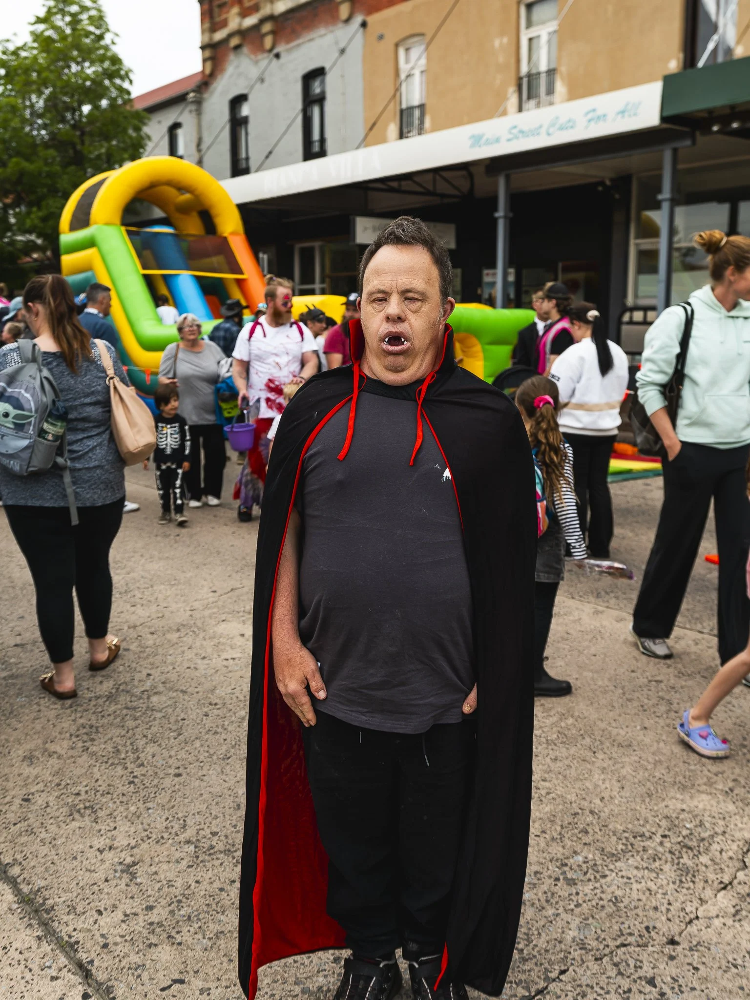 Man dressed as a vampire with a black cape and gray shirt at a crowded outdoor festival, with an inflatable slide and people in the background.