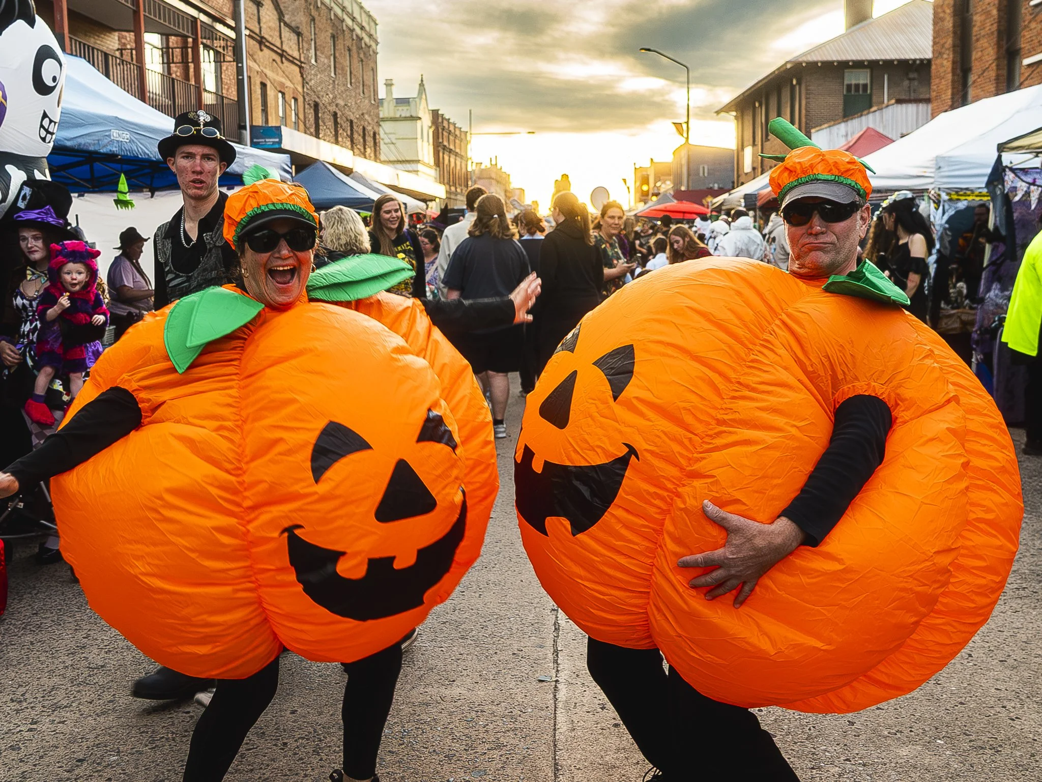 Two people dressed in inflatable pumpkin costumes with jack-o'-lantern faces, participating in a Halloween parade or festival on a city street at sunset, surrounded by many other festival-goers and vendor tents.