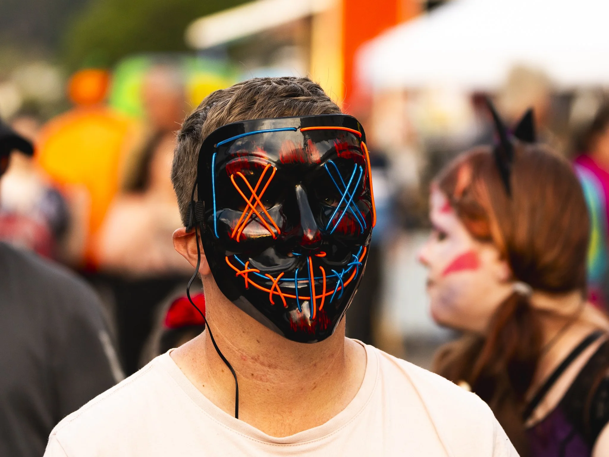 A person wearing a black mask with neon red and blue design, resembling a skull, at a crowded outdoor event.