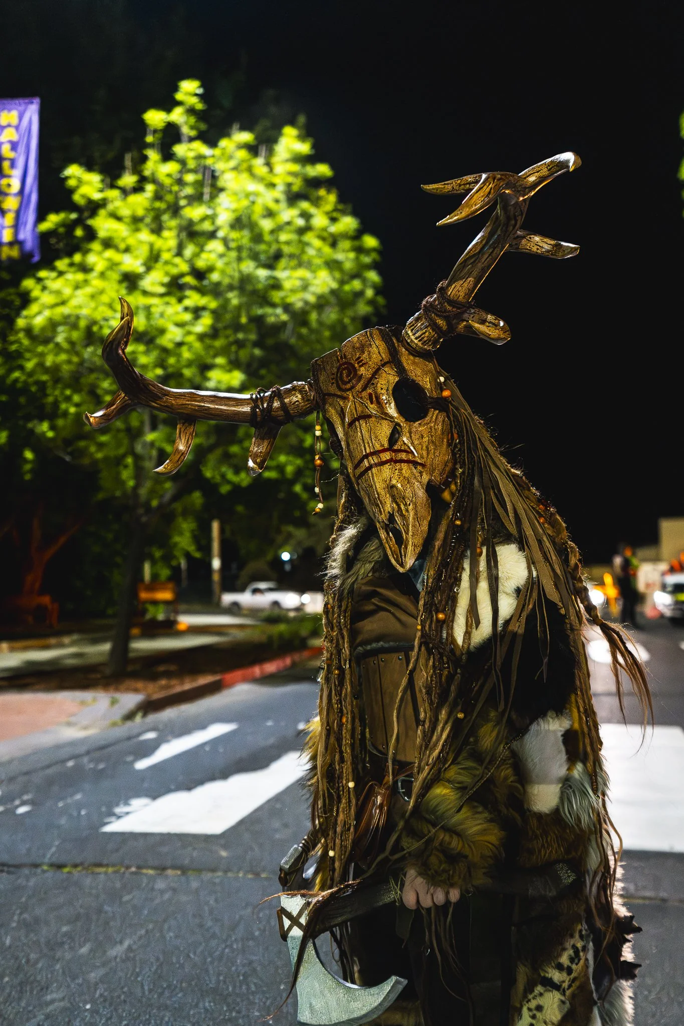 Person dressed in a costume with a wooden mask and antler headdress, holding a axe, nighttime outdoor setting.