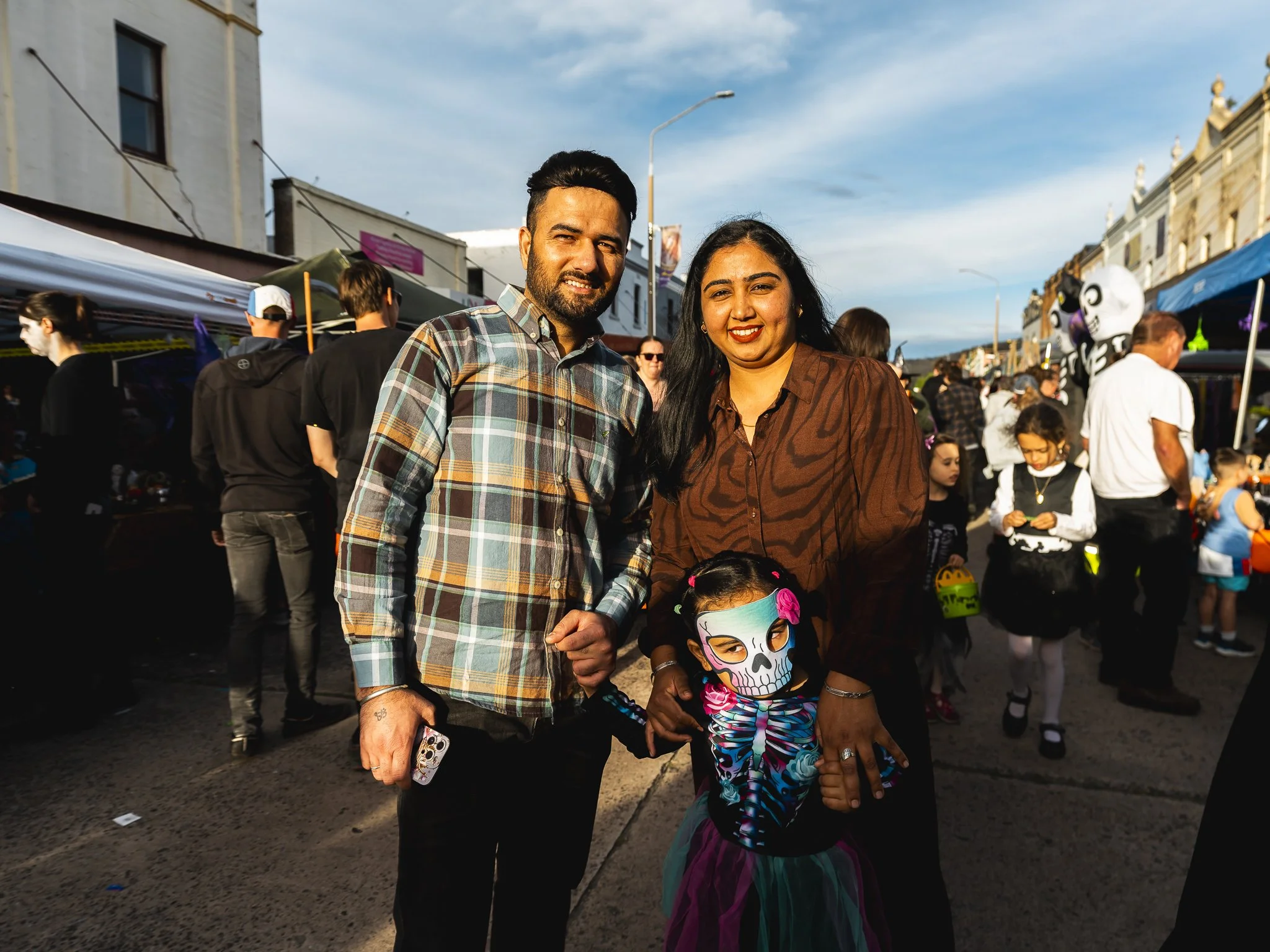 A family at a street fair dressed in costumes, with a man, a woman, and a girl wearing a skull mask.