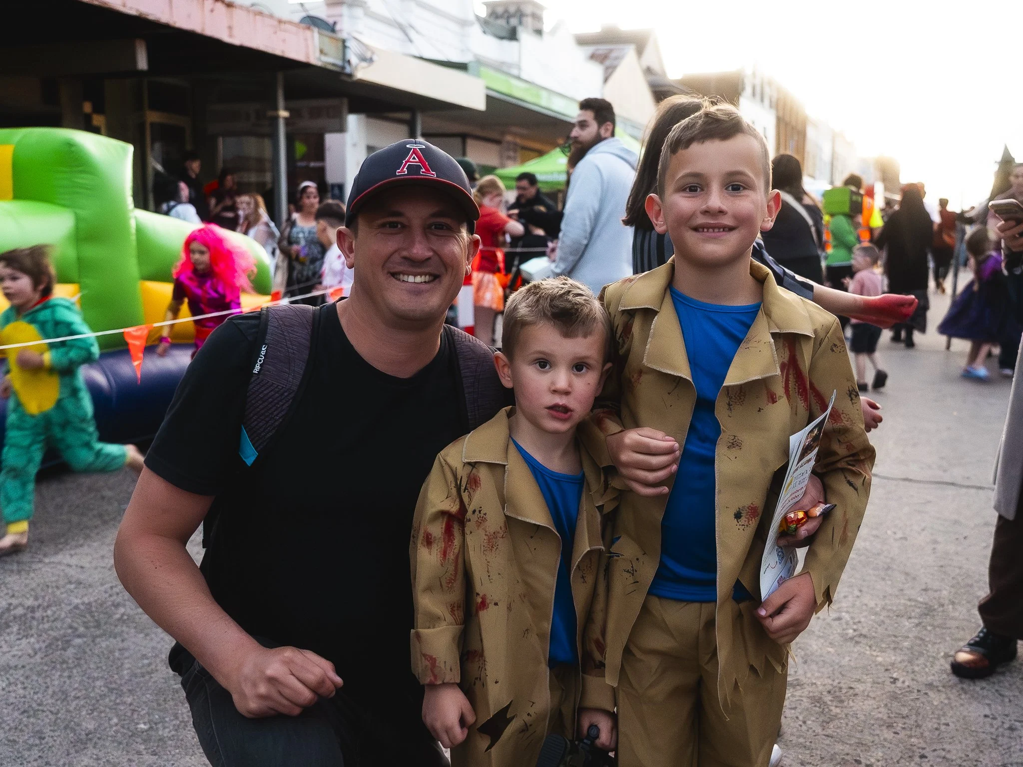 A group of three people, an adult man and two young boys, smiling at the camera at a busy outdoor event or festival, with other people and attractions visible in the background.