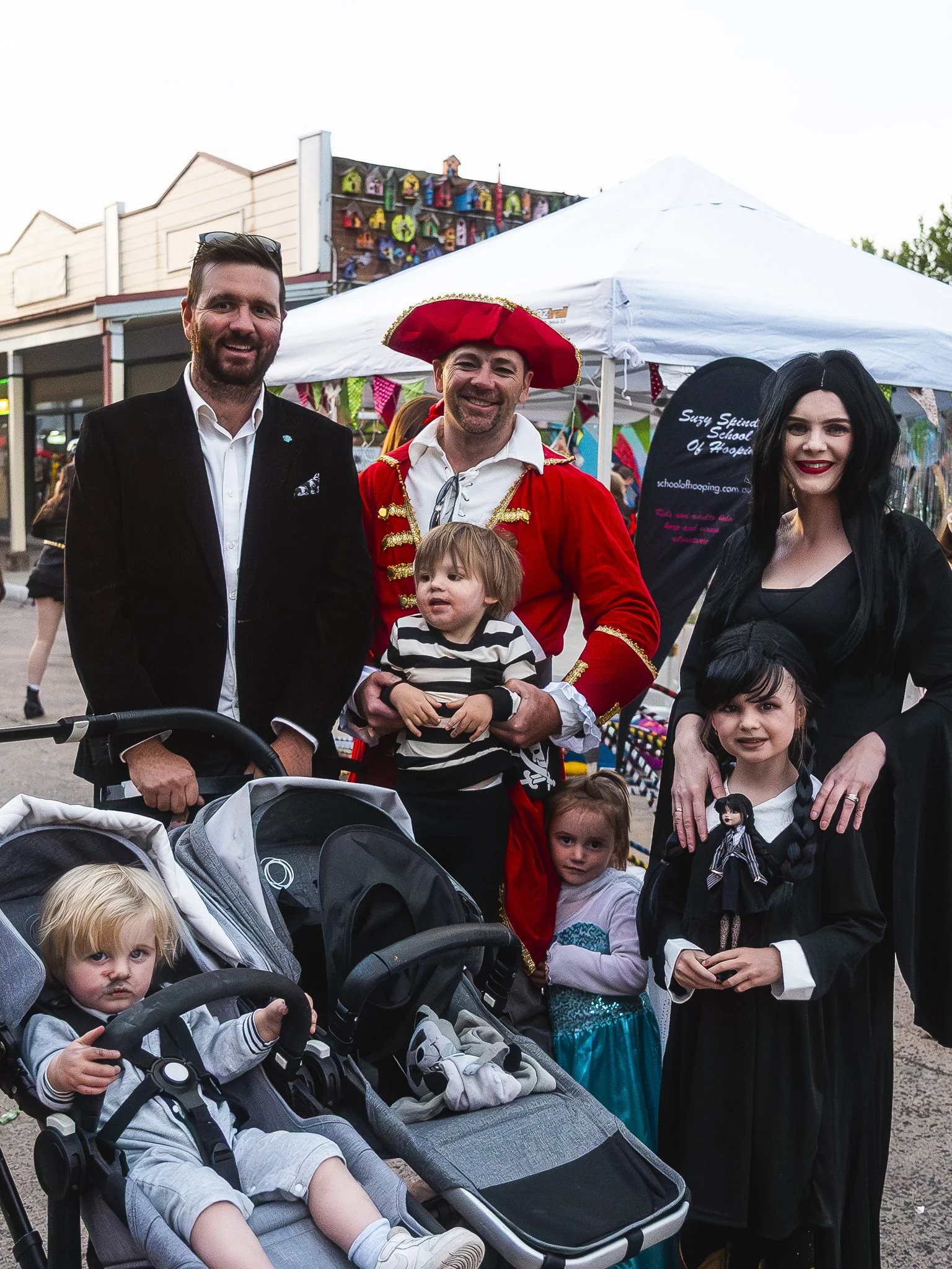A group of people dressed in costumes at an outdoor event. There are two adults in formal or costume attire, one man in an 18th-century style red coat and hat, and a woman with long black hair in a black dress. Three children are in the group, one in