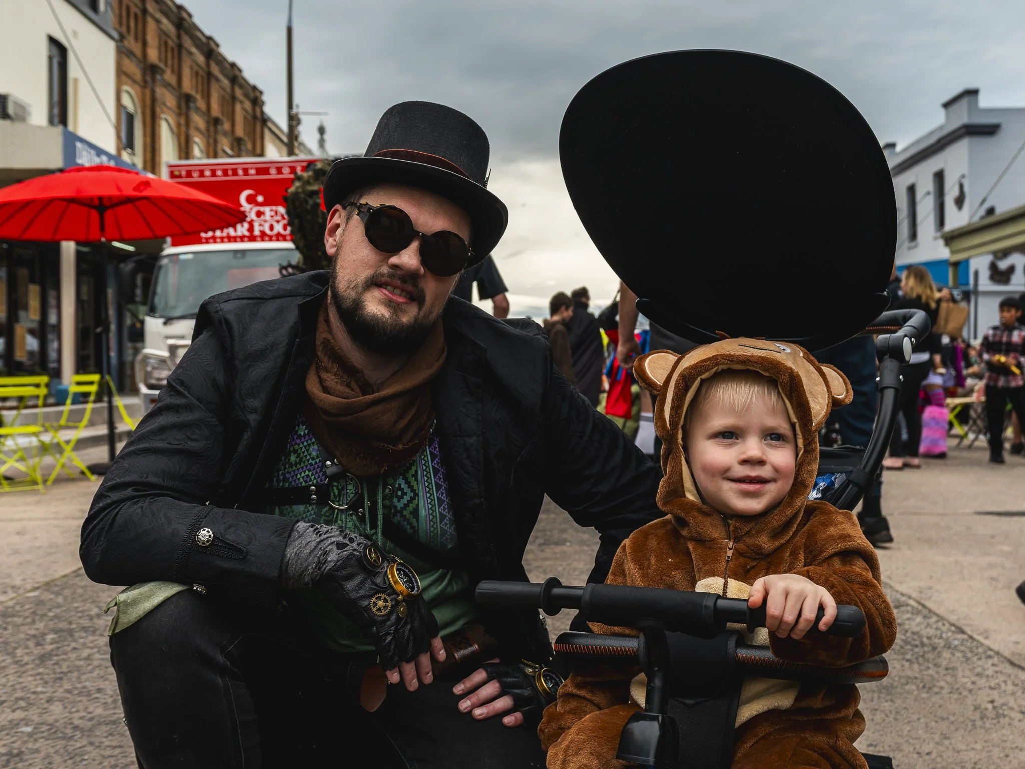 A man with a beard, wearing a black top hat, sunglasses, and a black leather jacket crouches next to a young child in a bear costume sitting in a stroller. They are at an outdoor event with other people, colorful umbrellas, and buildings in the backg