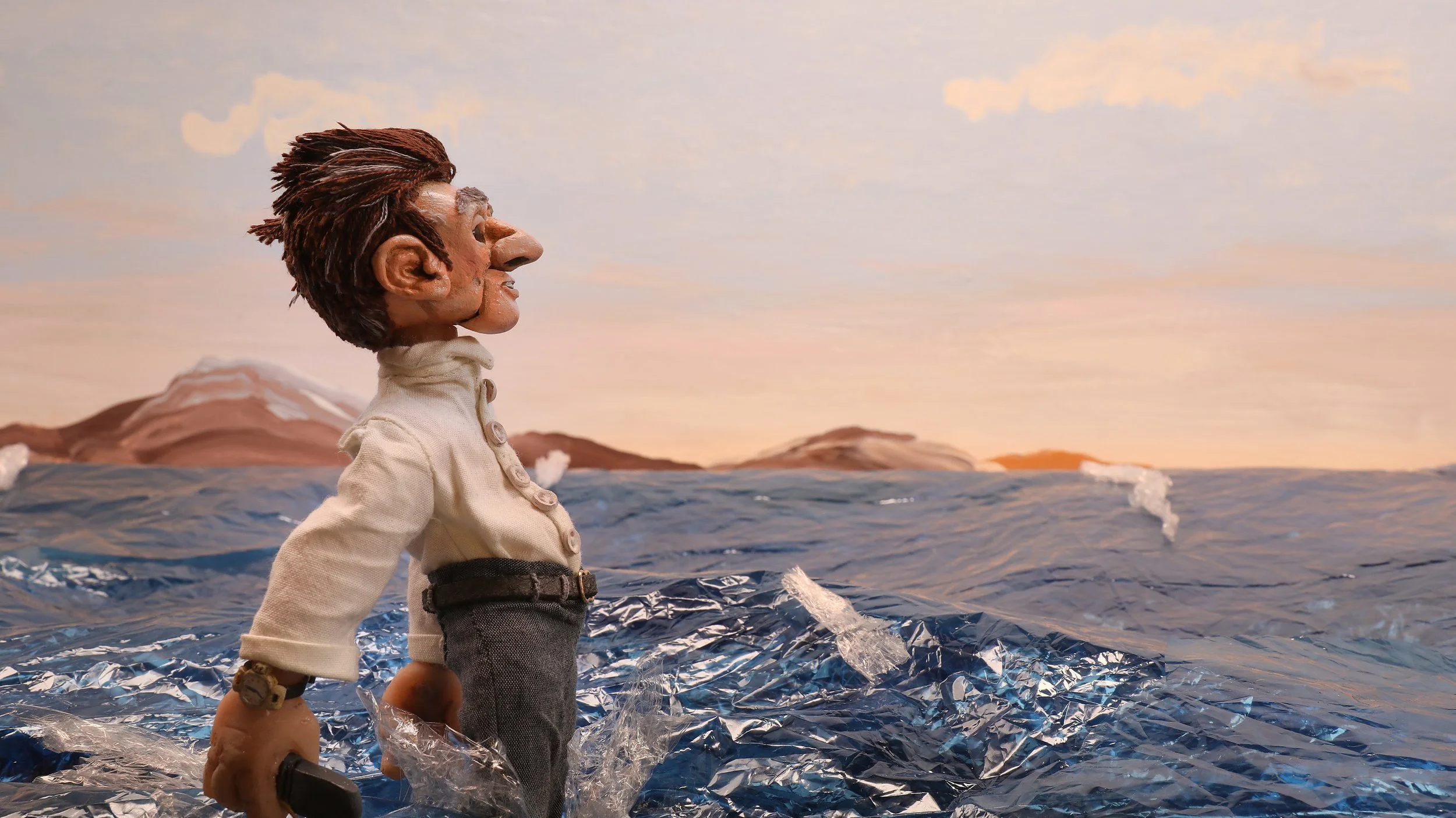 A puppet with brown hair, a white button up long-sleeved shirt, and grey trousers standing on a simulated ocean surface made of crumpled metallic material with mountains and a sunset sky painted in the background. Still from Cnut short film.