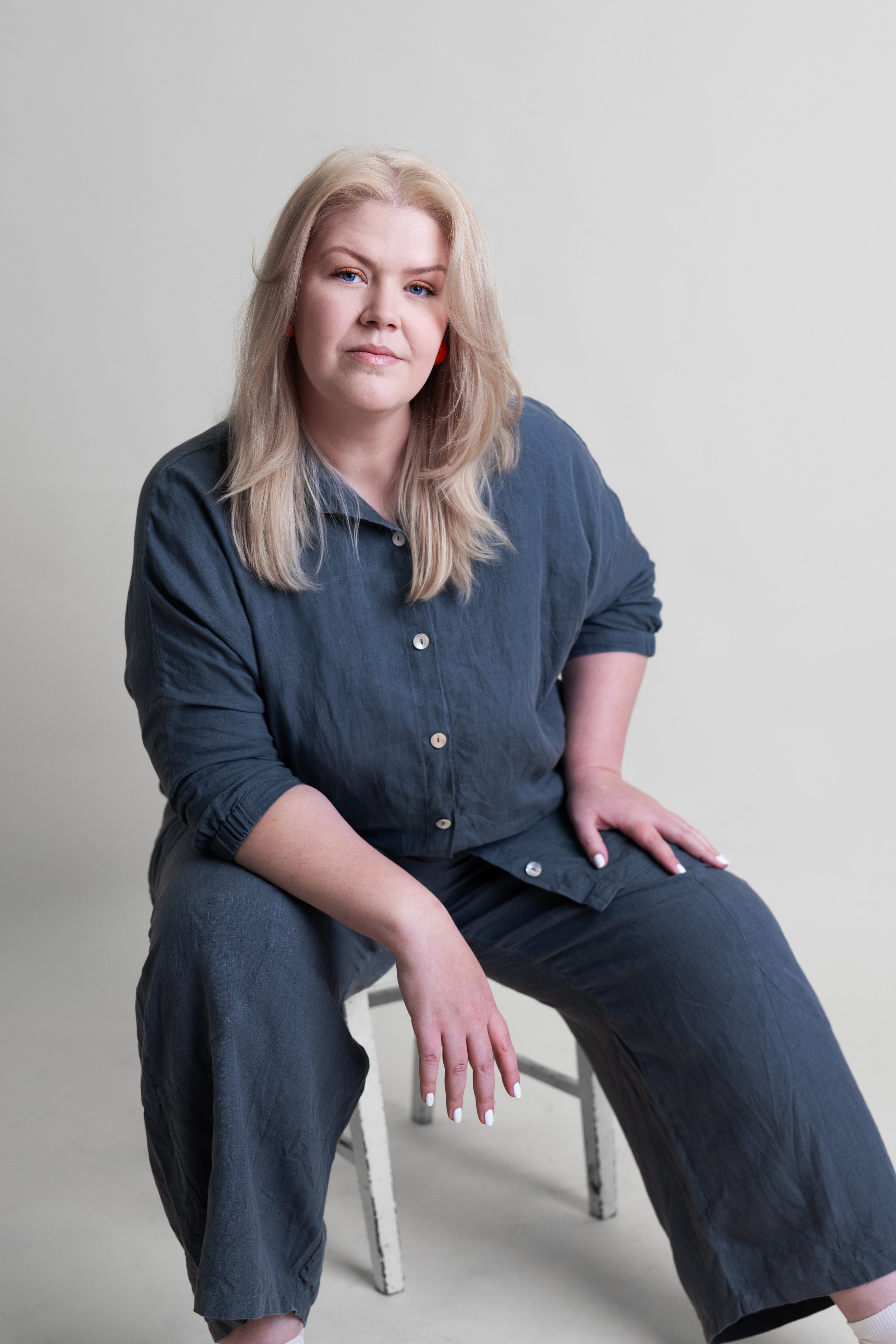 A woman (Courtney Westbrook) with long blonde hair sitting on a white stool against a plain light-coloured background, wearing a dark charcoal, blue button-up linen shirt and pants. Headshot style photograph.