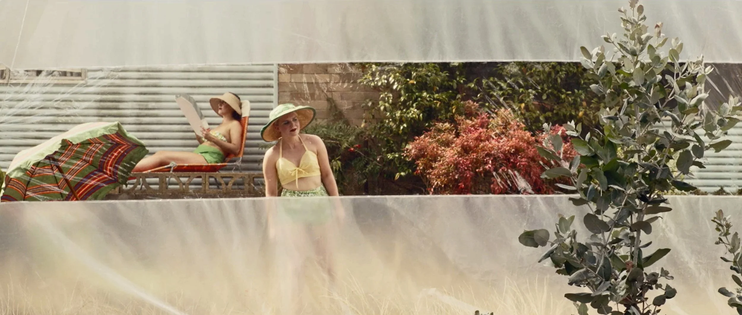 Two women in vintage swimsuits and sun hats relaxing outdoors, one on a lounge chair reading, the other standing, with a patterned umbrella in the foreground. They are in an enclosure. Film still from The End, The Beginning short film.