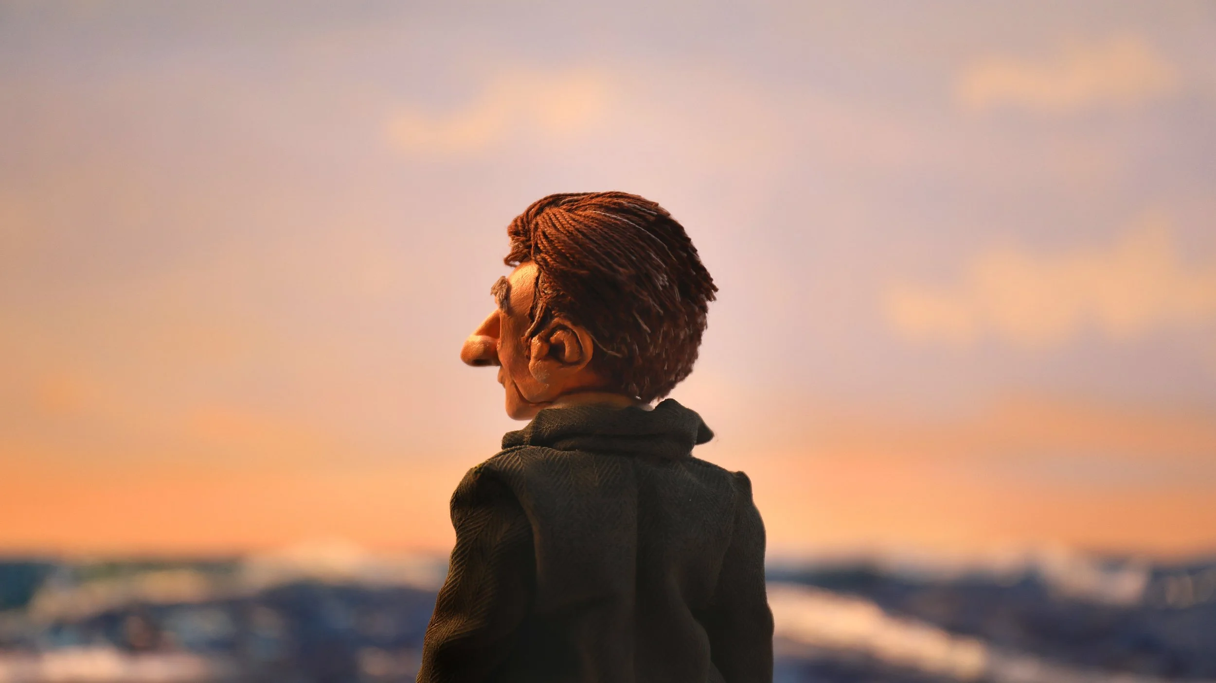 A figurine puppet of a man with brown hair in a slicked-back style, wearing a dark jacket, stands facing away at sunset over ocean, with the sky painted in soft pastel hues and the ocean in the background. Film still from Cnut short film.