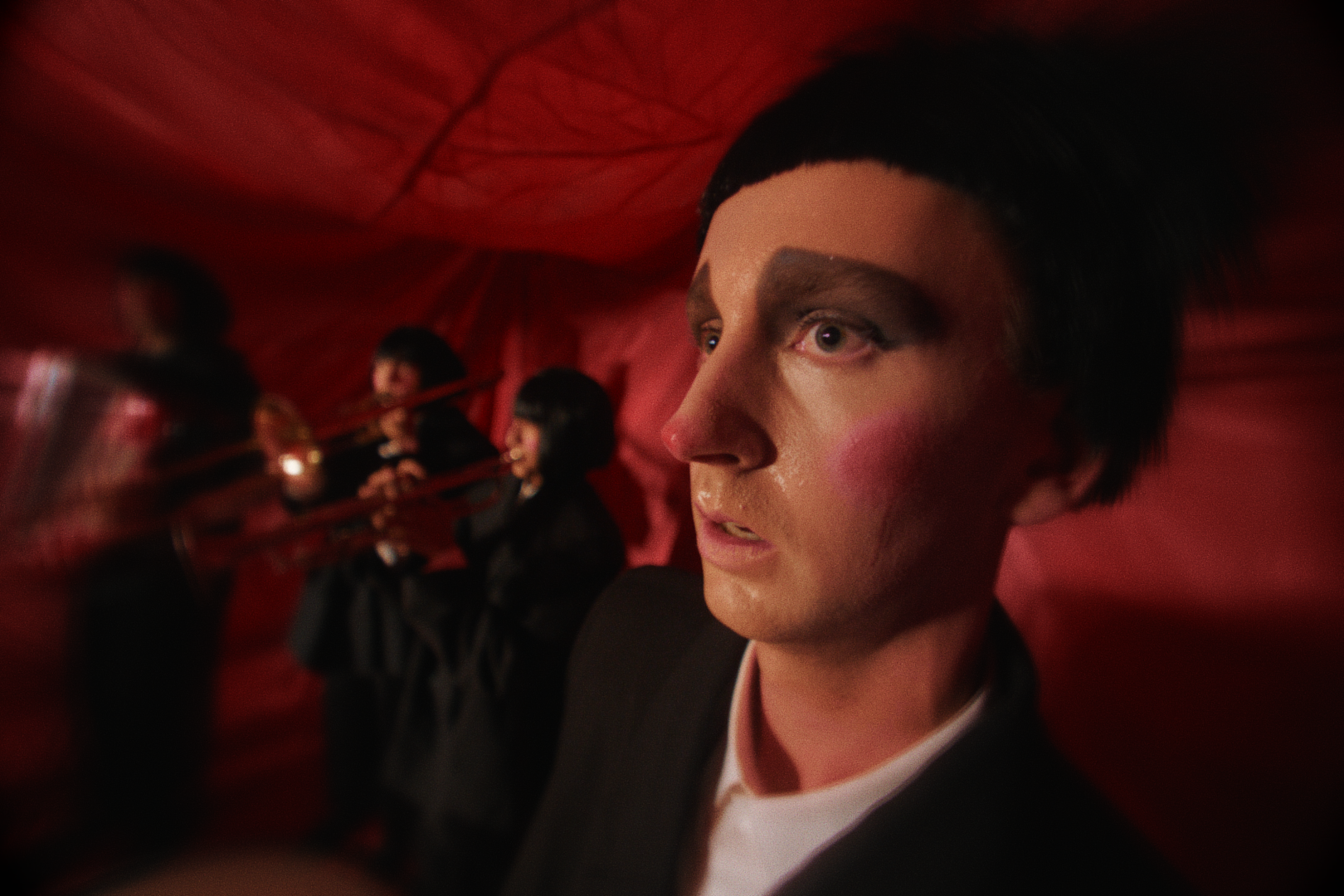 Close-up of a man with dark hair, red eyeshadow, wearing a suit, looking to the right, with a blurred background of women in black outfits playing trumpets against a red backdrop. Film still from Luna and the Brain Tuna short film.