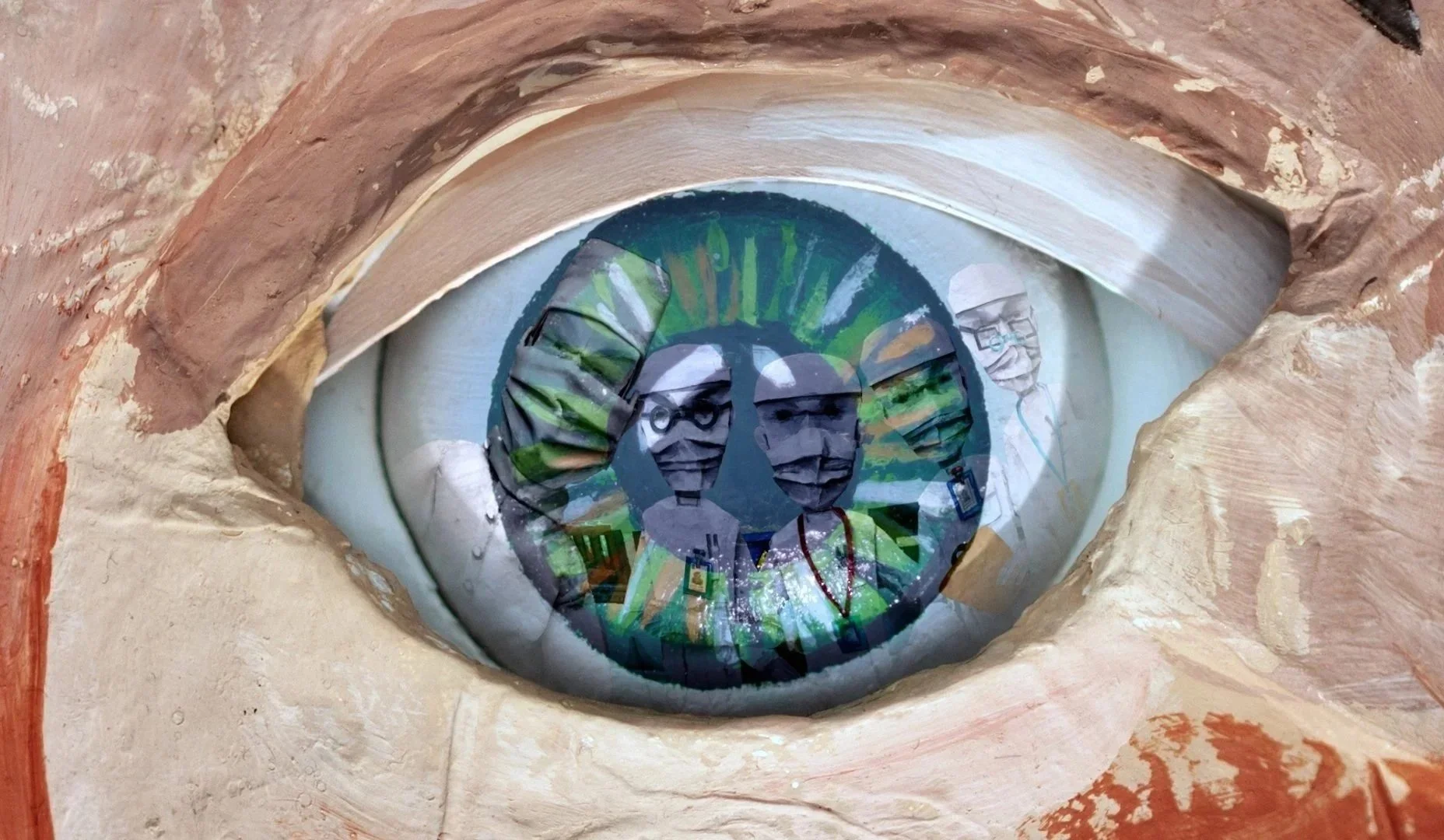 A close-up view of a green eye from a paper puppet, surrounded by textured skin with layers of paint. The eye features a reflection of five doctor figures, including two with glasses and a third with a lanyard. Film still from If/When short film.