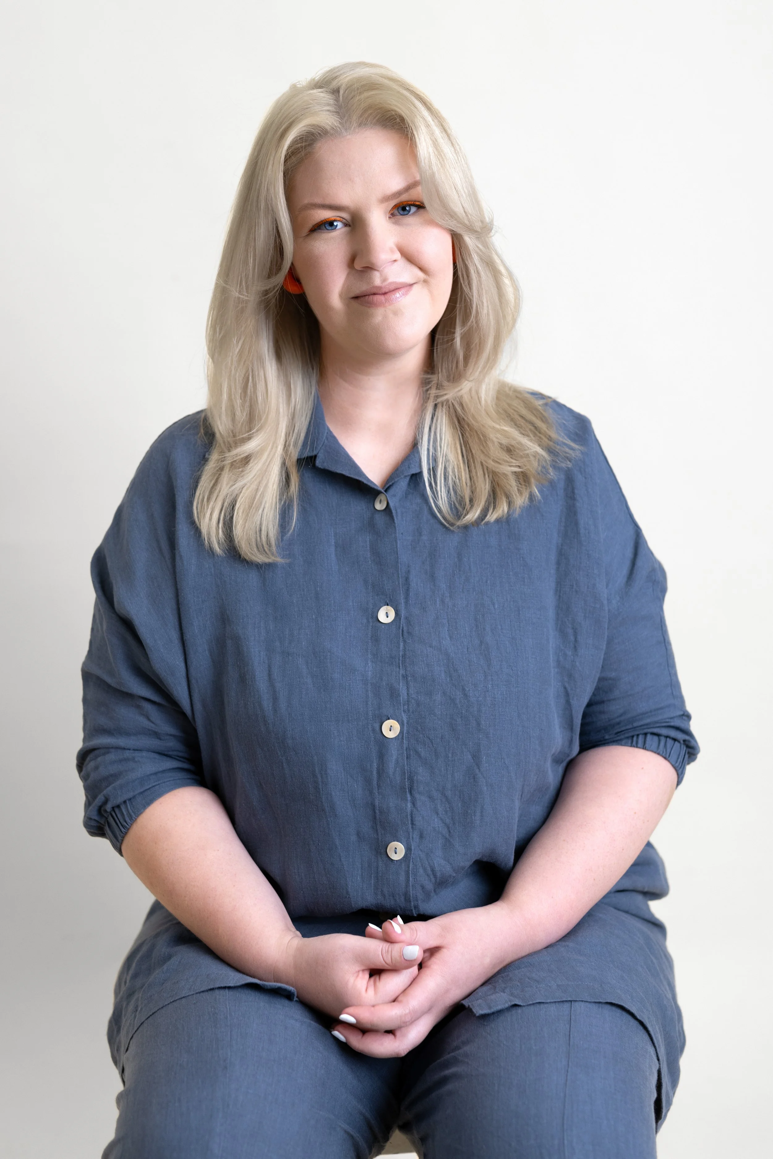 A woman (Courtney Westbrook) with blonde hair, blue eyes, and orange earrings sitting against a plain light grey background, wearing a blue charcoal button-up shirt and pants. A headshot style image.