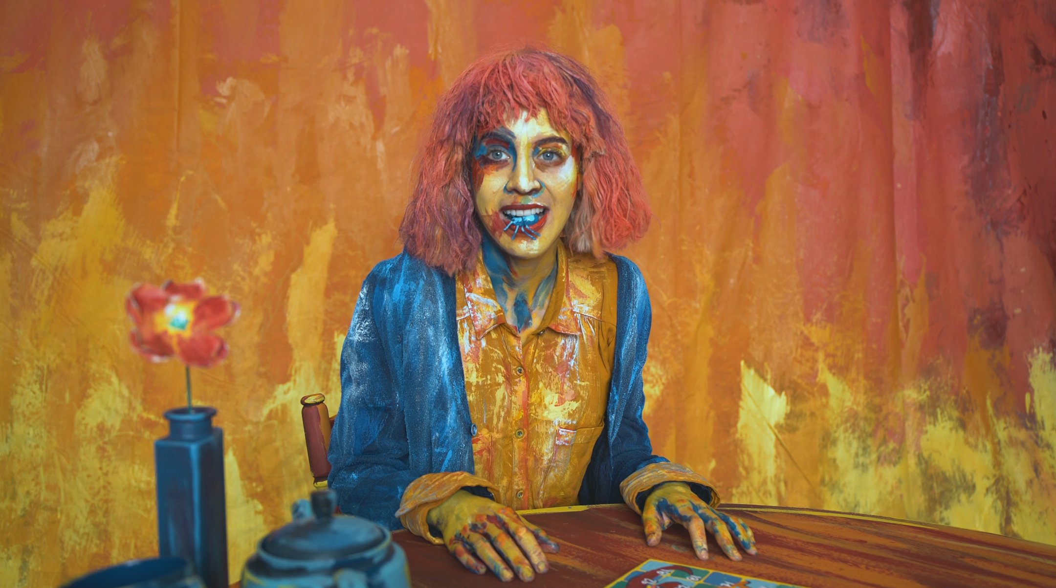 A woman with multi-coloured painted face and hair, dressed in a blue and yellow jacket and shirt, sitting at a table with a colourful background. The table has a vase and flower. All hand painted. Film still from Theo & Celeste short film.
