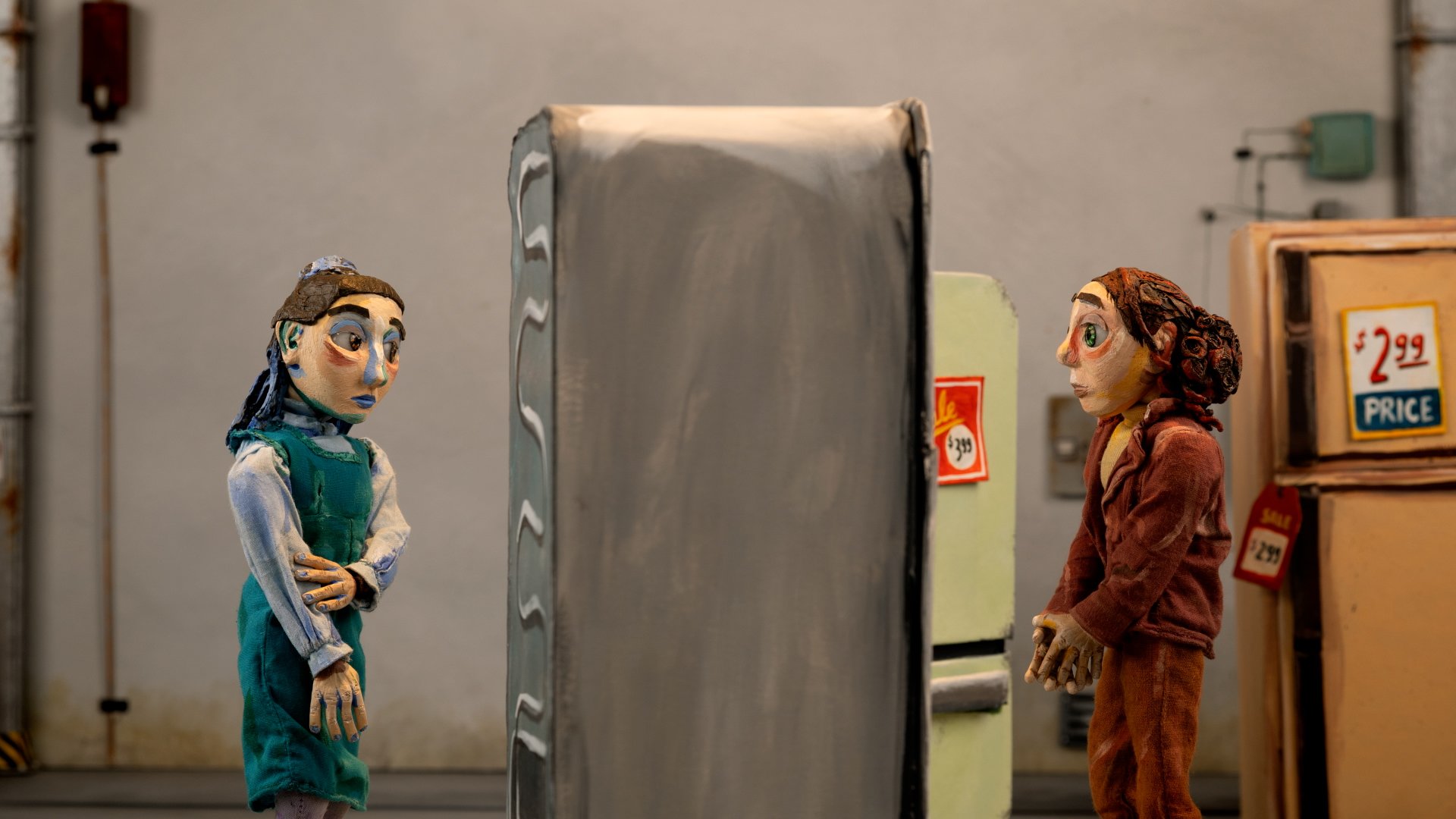 Two puppets with human-like features are in a fridge store, facing each other. The puppet on the left is dressed in a green dress with light blue sleeves, the other in a brown, orange outfit with curly hair. Film still from If/When short.