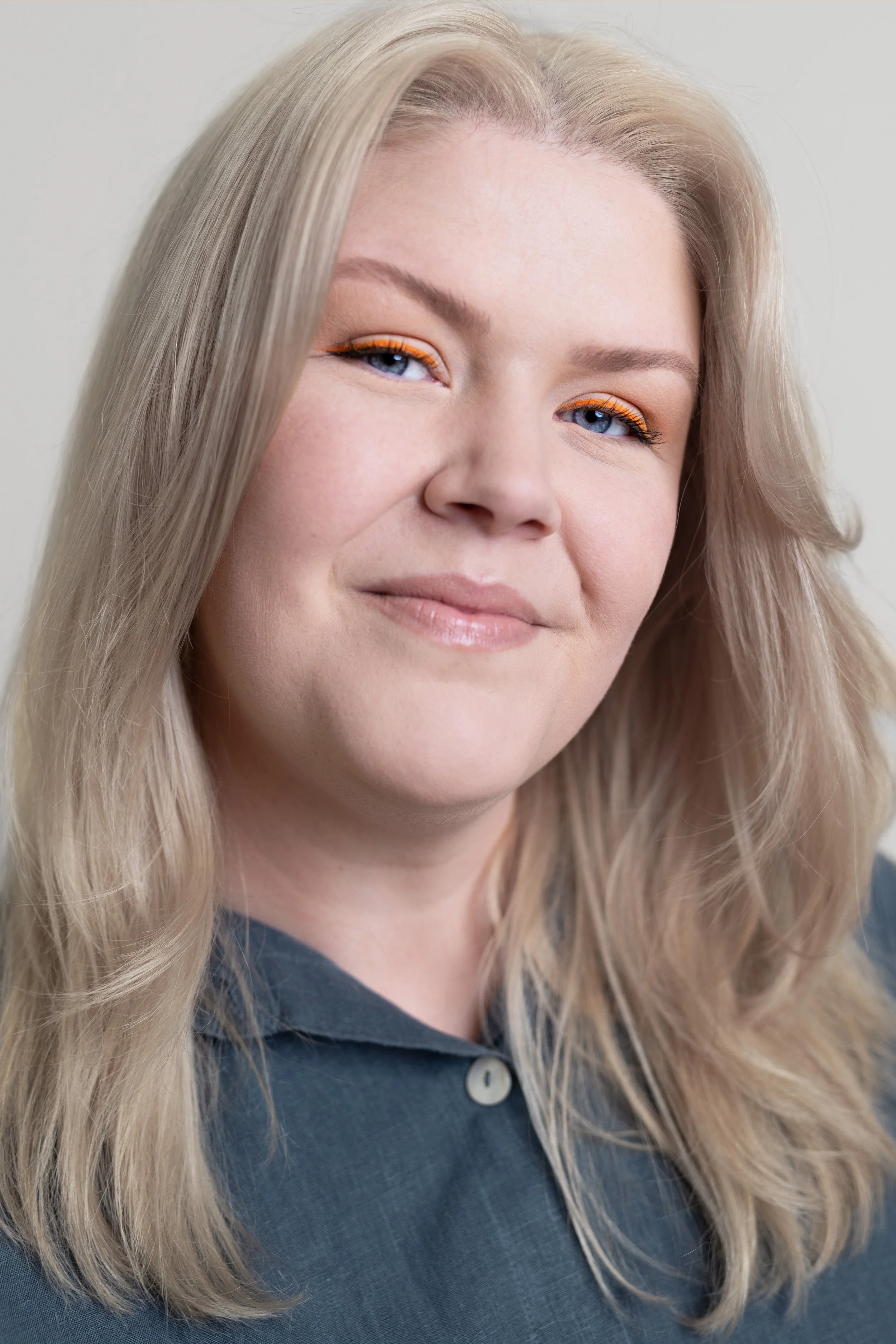 A close-up photograph of a woman (Courtney Westbrook) with long blonde hair, light blue eyes, and makeup including orange eyeliner, wearing a dark-coloured charcoal linen shirt with a buttoned collar, against a plain light background.