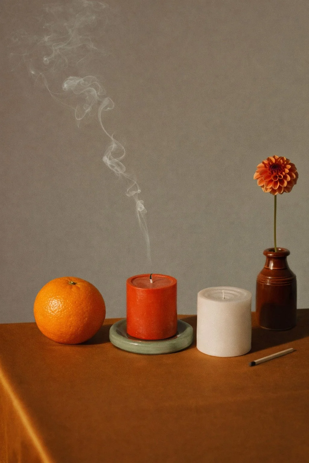 An orange fruit, a vibrant orange pillar candle with smoke rising. The orange candle is on a sage green ceramic plate. A white candle, and a small orange flower in a brown vase on a table surface covered in rust colour silk.