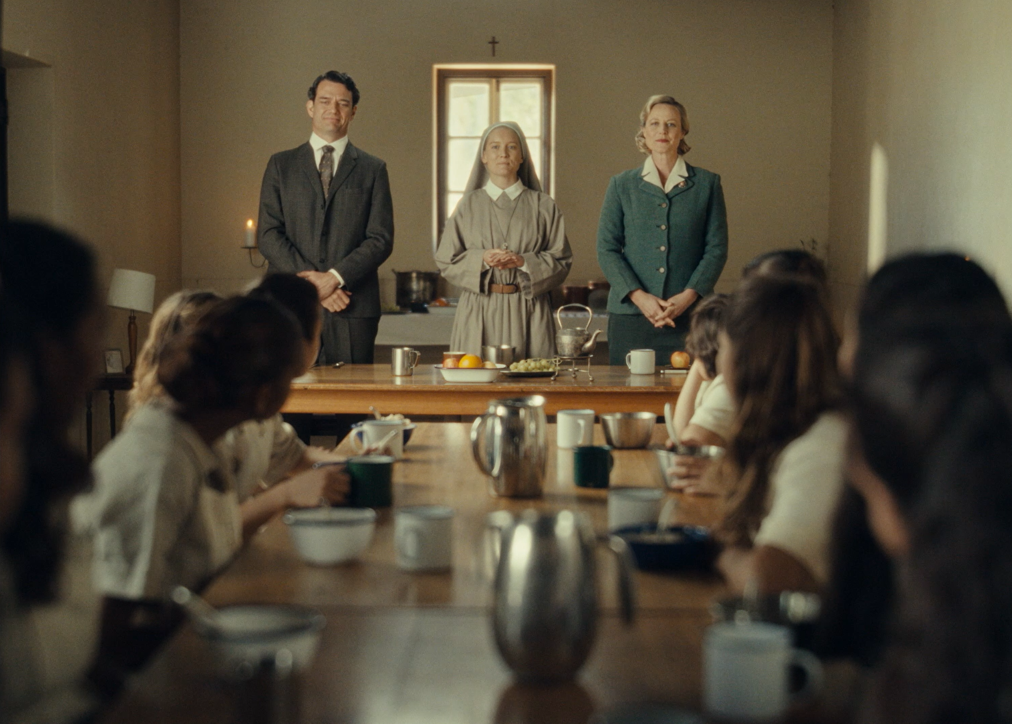 A scene from a historical or period film showing three people standing in front of a window with a cross above it, in a room with a table set with cups, bowls, and food, while several children sit at the table. Australian short film Finding Jedda.