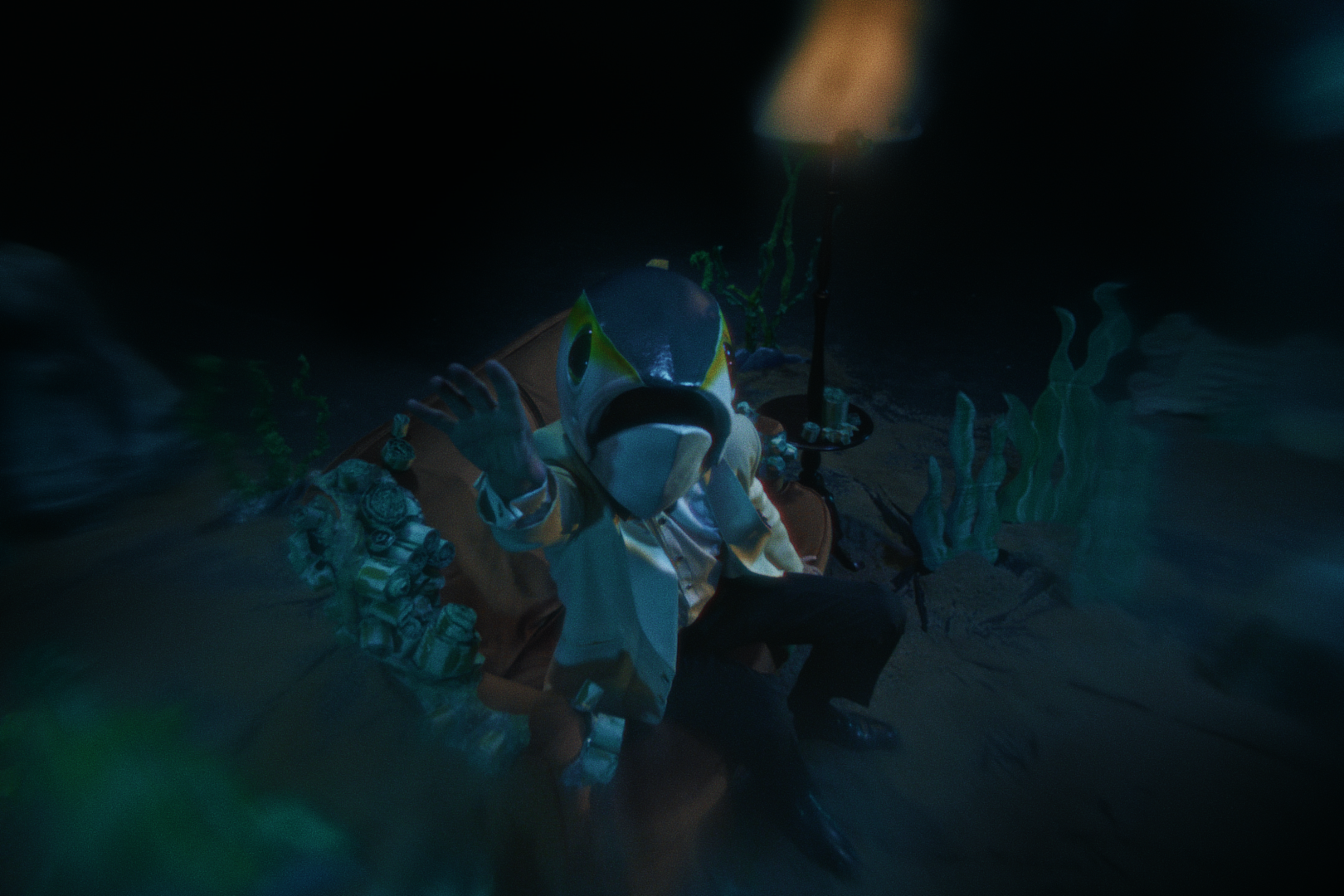 A man wearing a tuna-inspired puppet head mask and a white suit costume, sitting on an orange couch that is in a dark underwater-themed setting with coral decorations and an industrial-style lamp. Film still from Luna and the Brain Tuna short.