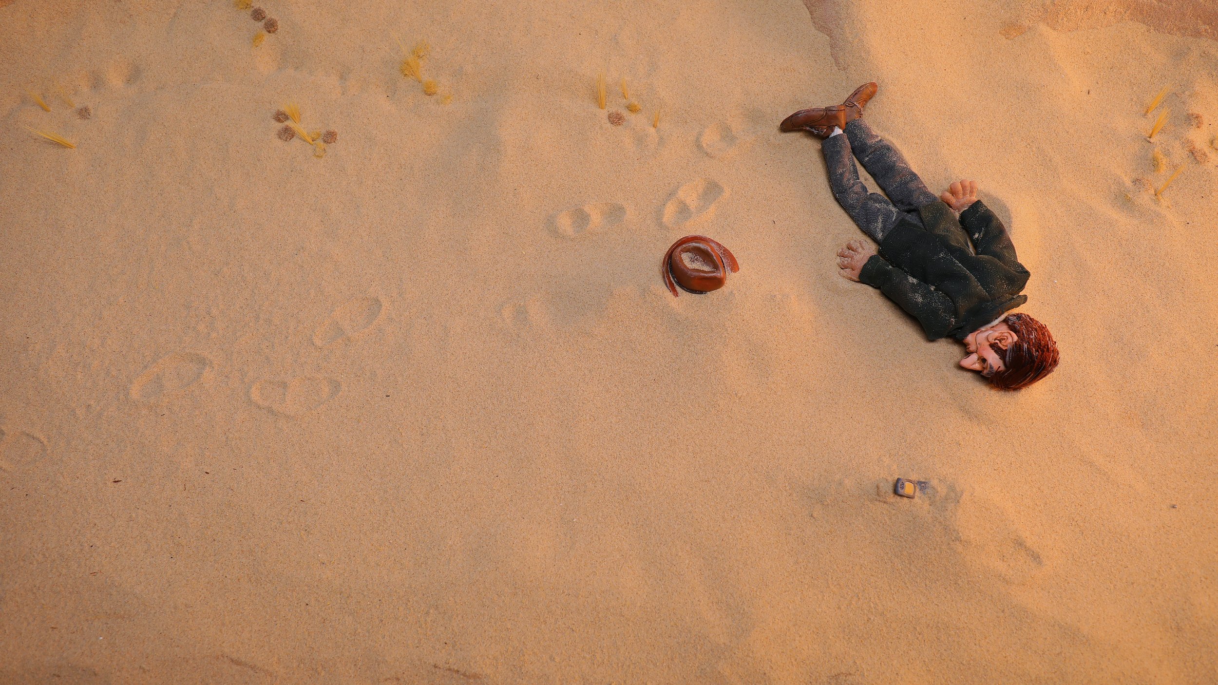 A man puppet lying face-down on a sandy surface, wearing a dark green jacket, trousers, and brown leather shoes. Their head is turned to the side with brown hair visible. There is a small hat and a phone near them on the sand. Film Still from Cnut.