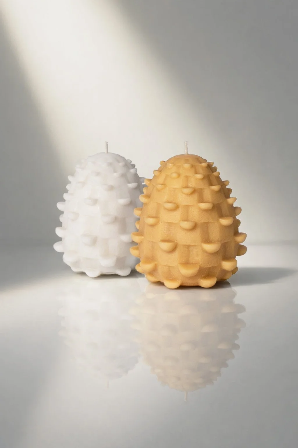 Two decorative candles shaped like pinecones, one white and one beige, sit on a reflective surface against a light background.