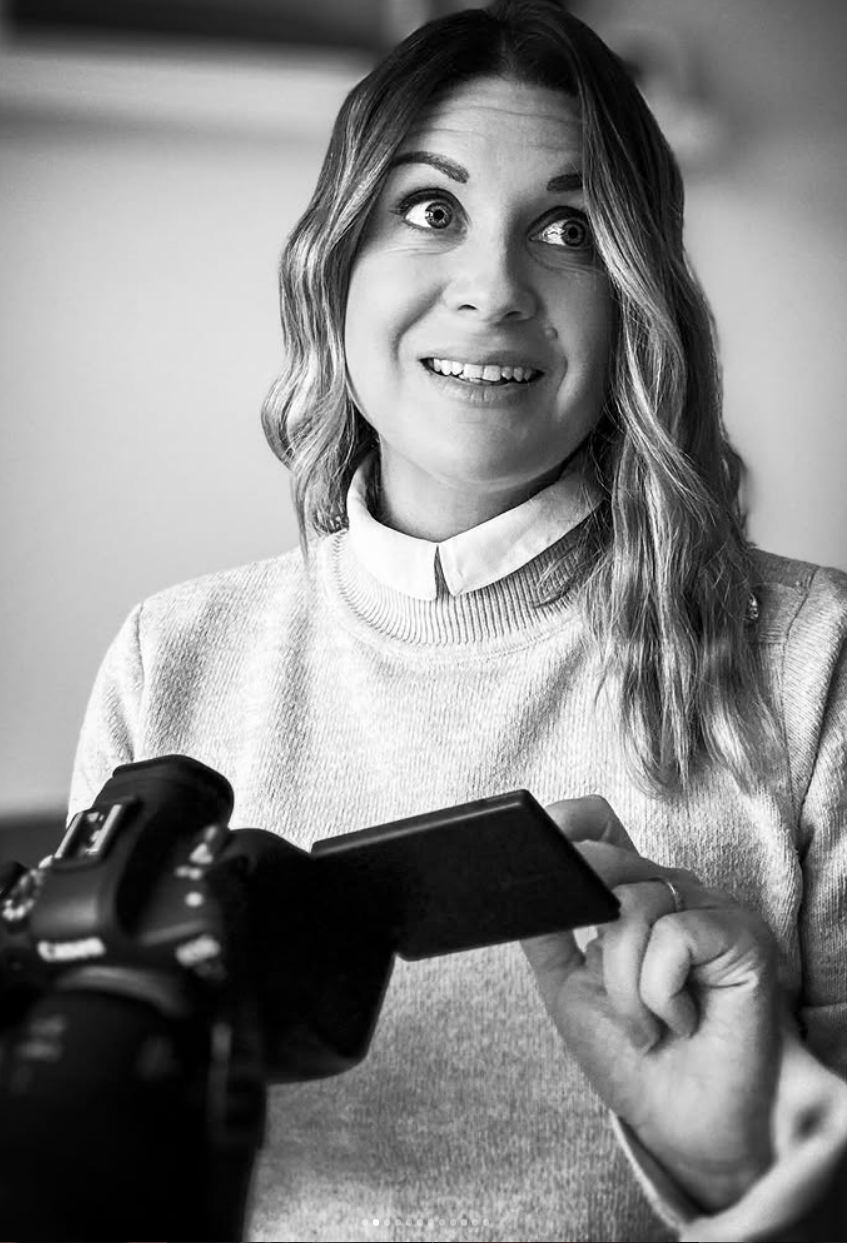 Une femme souriante tient un appareil photo et regarde la caméra. Elle porte un pull et une chemise à col. L'image est en noir et blanc.