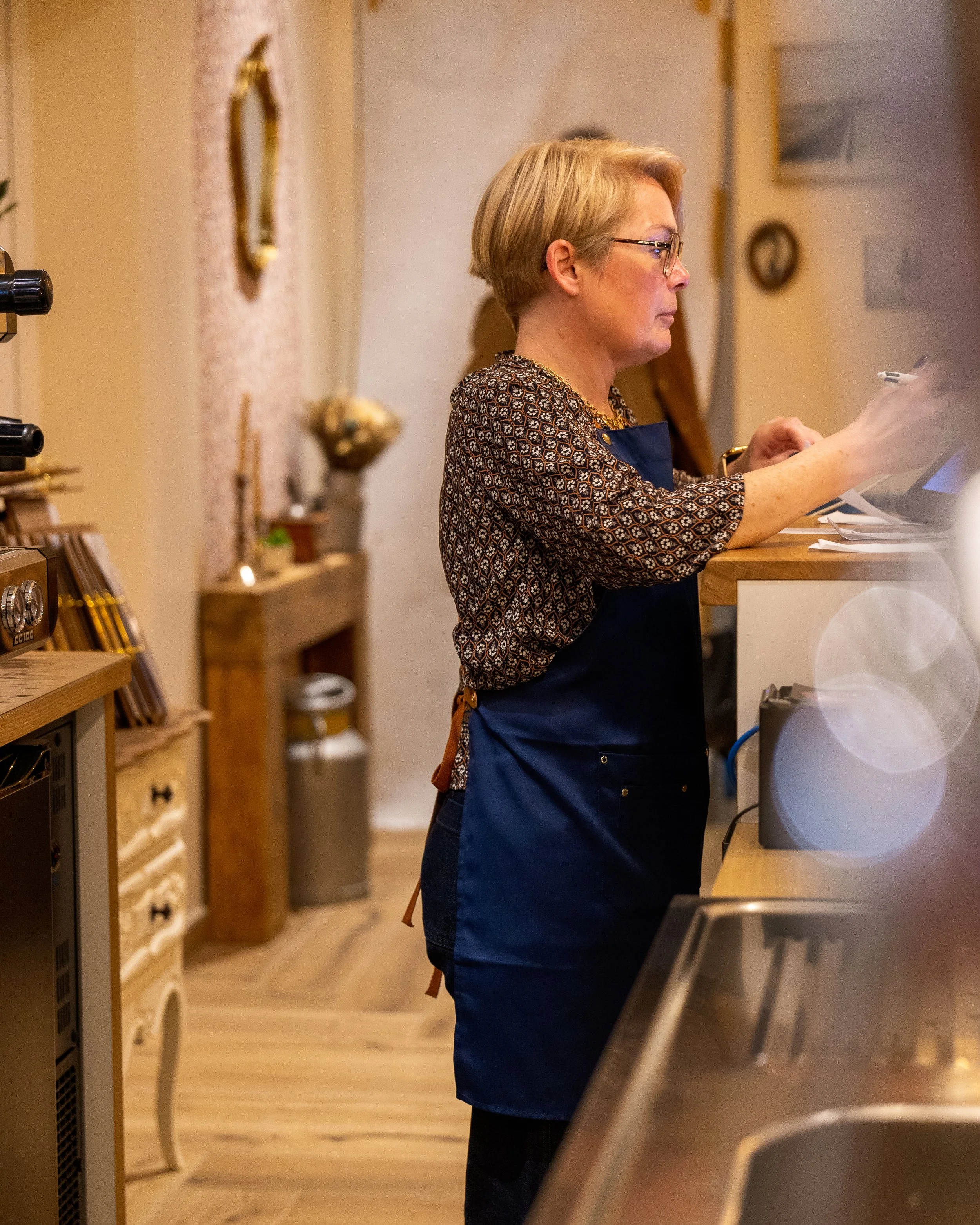 Une femme portant des lunettes et un tablier bleu travaille derrière un comptoir en bois, utilisant un appareil électronique dans une pièce décorée avec des cadres, des horloges et des objets artisanaux.