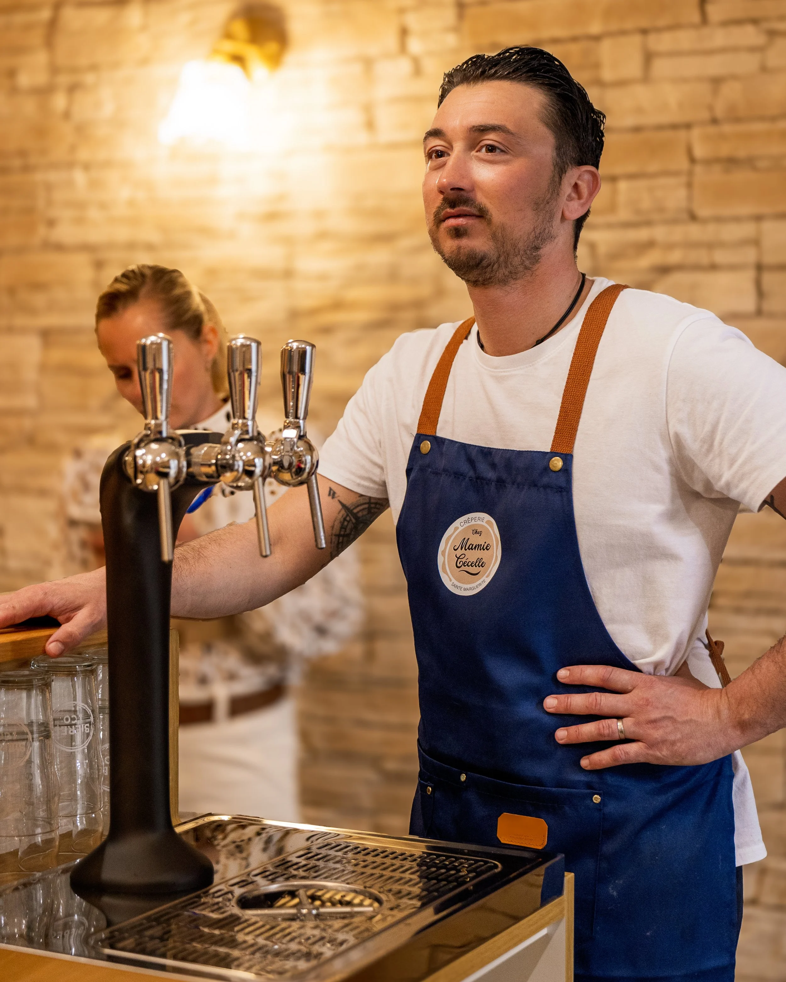 Homme portant un tablier bleu, debout derrière un bar en train de servir, avec une femme en arrière-plan dans un café ou un bar en bois.