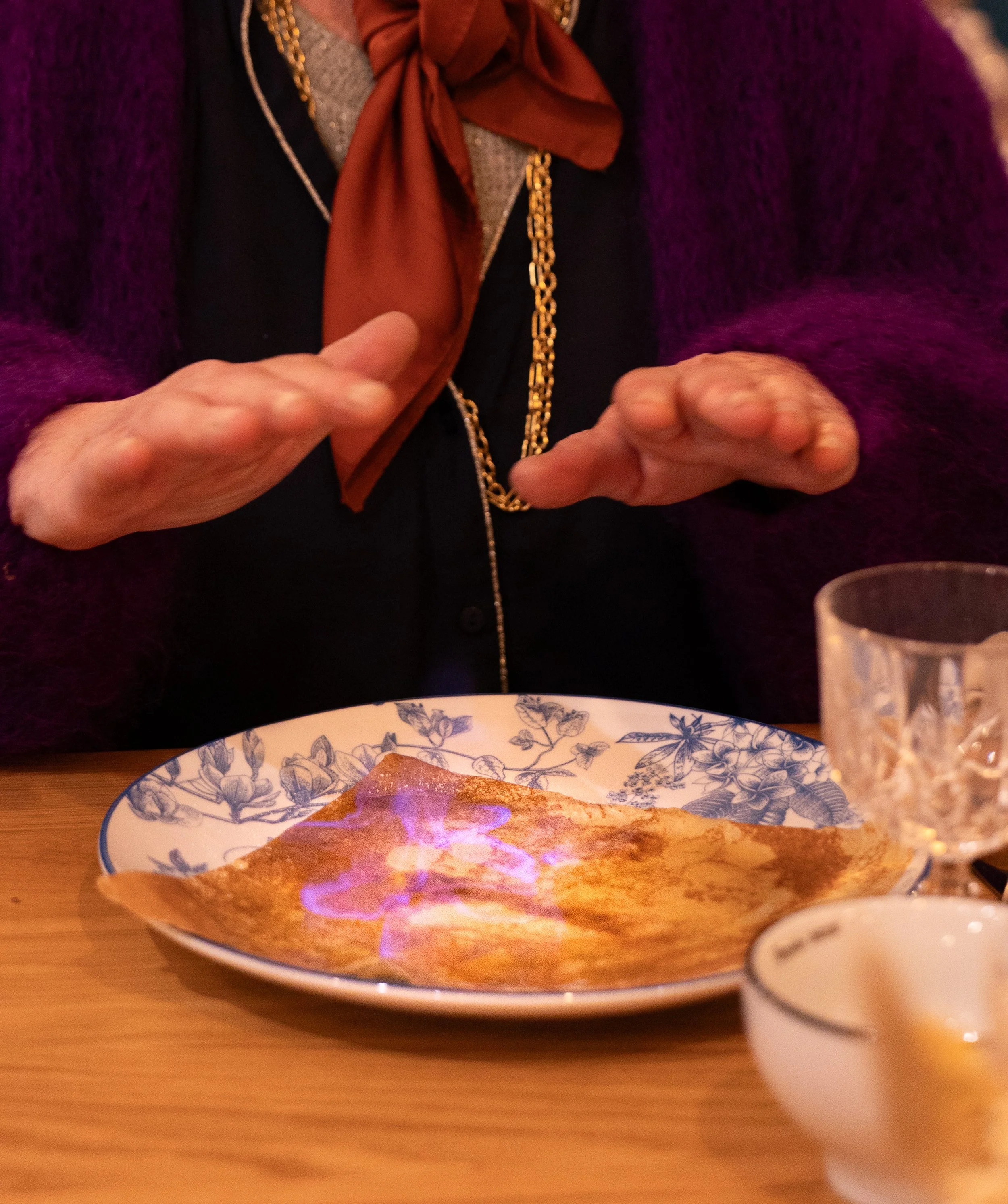 Une personne portant un foulard marron, une chaîne en or, et une veste violette est visible de la poitrine en haut. Elle semble être entrain de faire un geste avec ses mains. On voit aussi une assiette avec un plat, un verre à boisson, et une tasse sur la table.