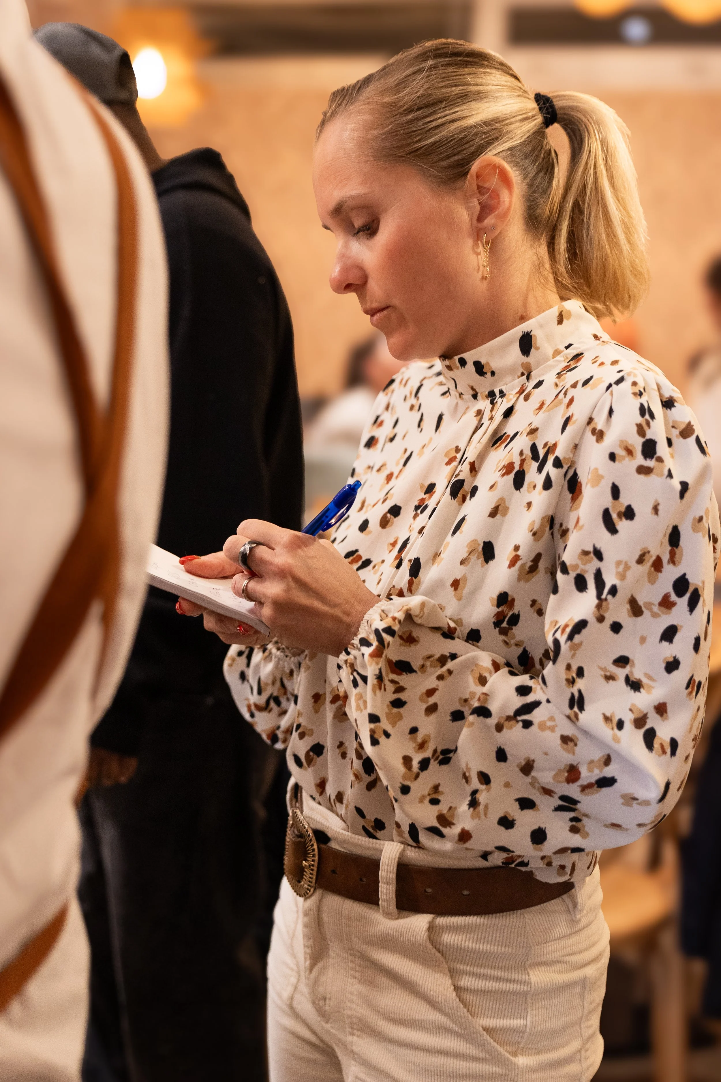 Une femme en haut d'un café ou restaurant, portant un chemisier à motifs avec un pantalon beige, tenant un stylo et un carnet, en train d'écrire ou de prendre des notes, dans une ambiance chaleureuse et conviviale.