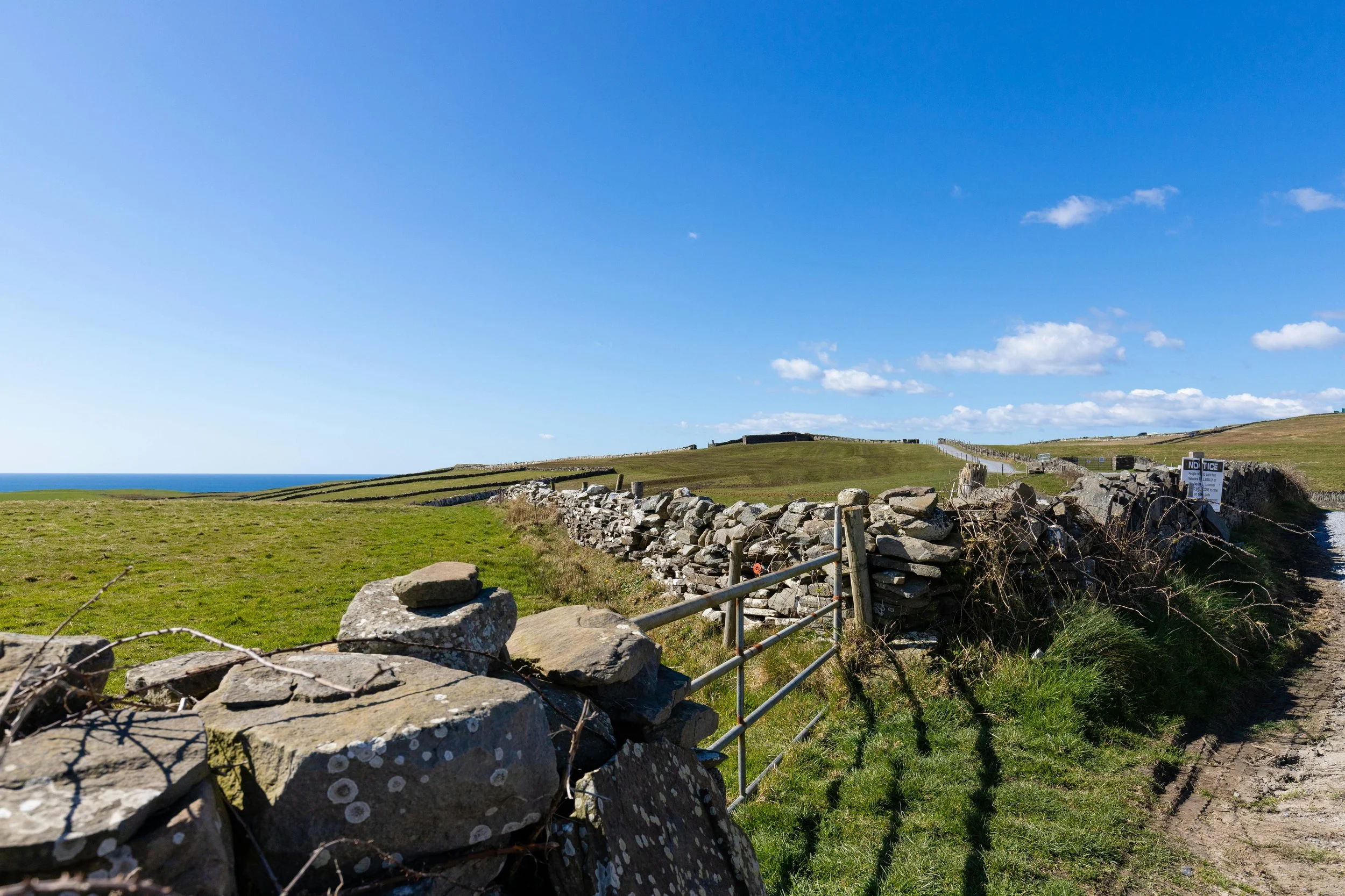 A scenic view of green grassy fields, stone walls, and a small fence under a bright blue sky with scattered clouds, with the coast and ocean visible in the distance.