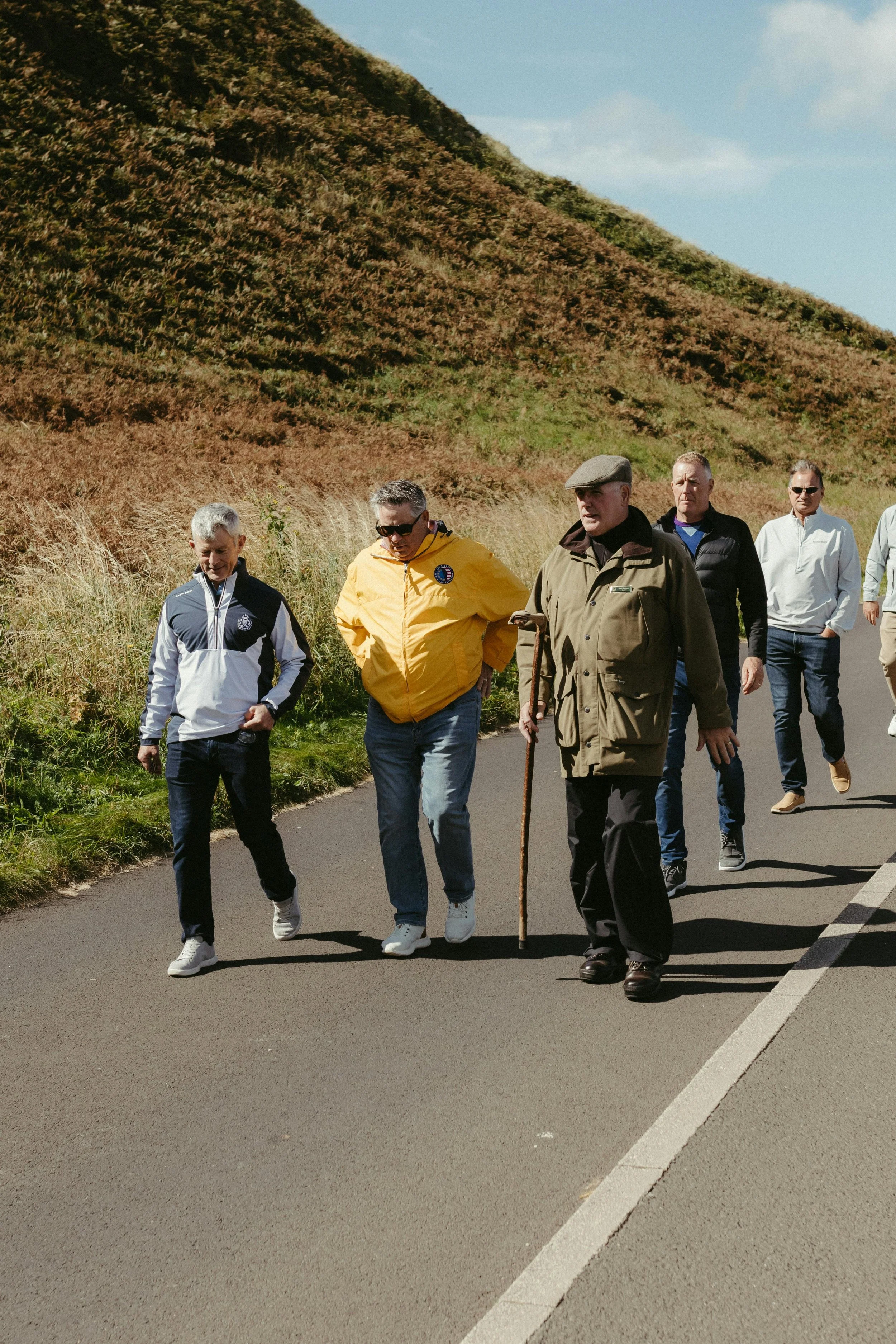 A group of six men walking along a paved path in a rural area with a grassy hillside in the background. They are dressed casually, with one man using a walking stick and another looking at his phone.