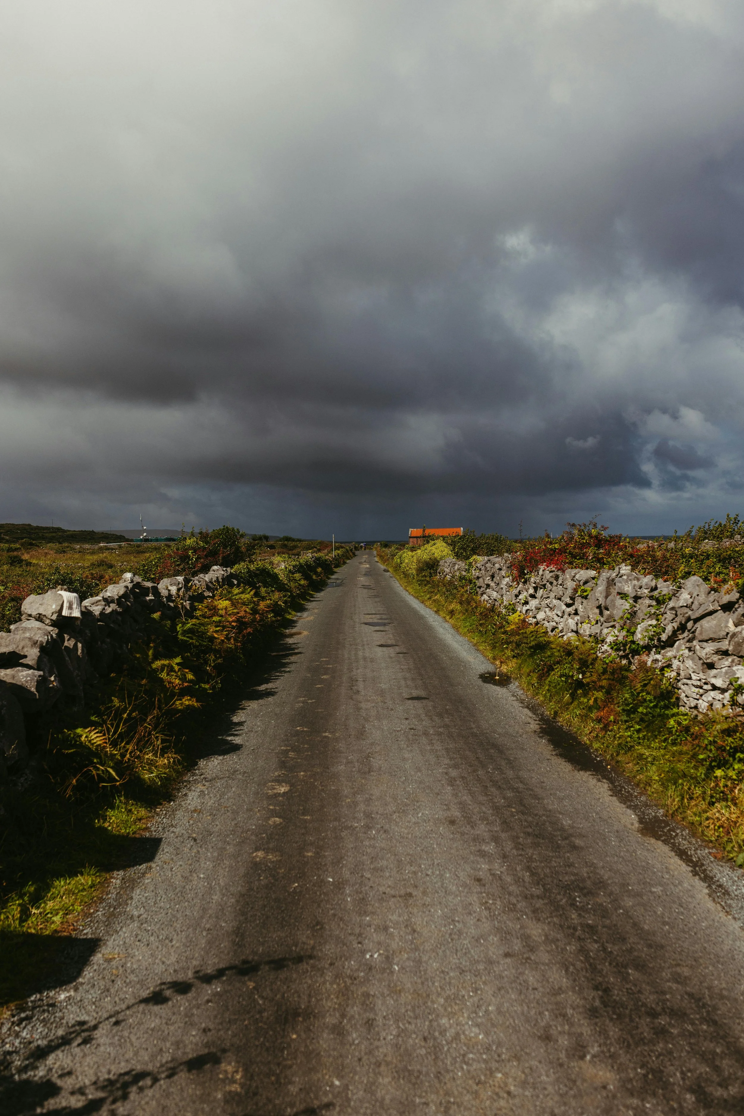A rural gravel road stretching into the distance, flanked by stone walls and green vegetation, under a dark, cloudy sky.