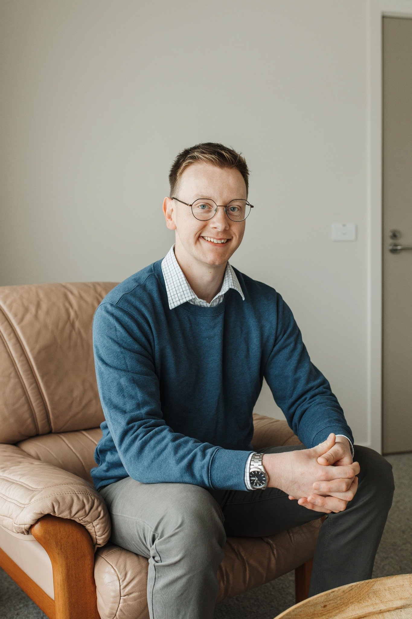 Matthew Eden, registered psychologist, seated on a sofa wearing glasses, a blue sweater and checked shirt.